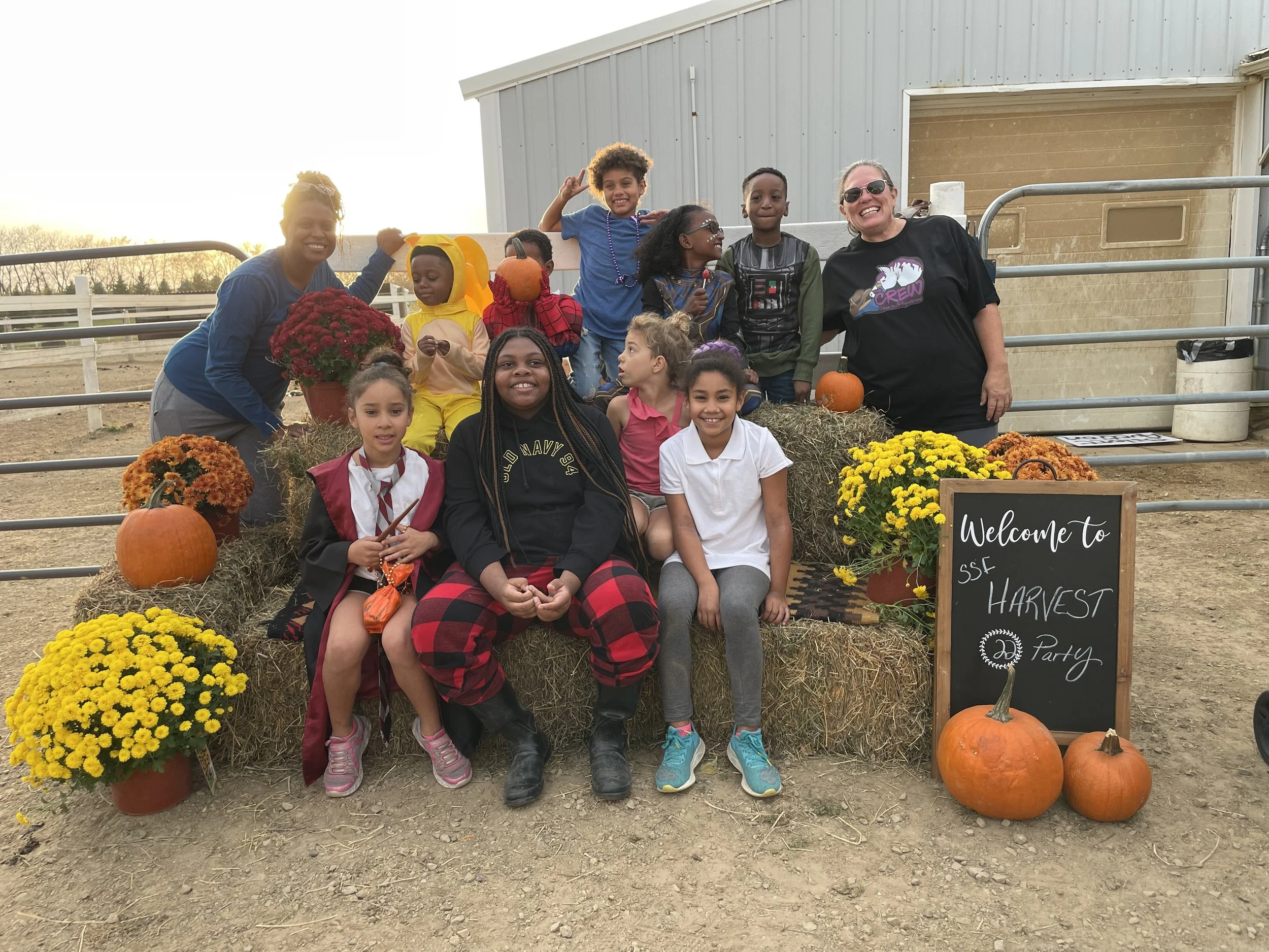 Group of children and adults at a harvest party, surrounded by pumpkins and flowers, with a chalkboard sign reading 'Welcome to SSF Harvest Party.'