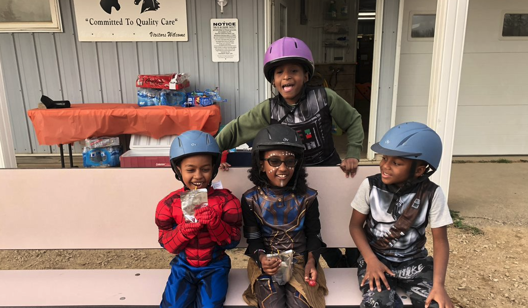 Children wearing costumes and helmets sitting on a bench, smiling; one dressed as Spider-Man, the others in various costumes.