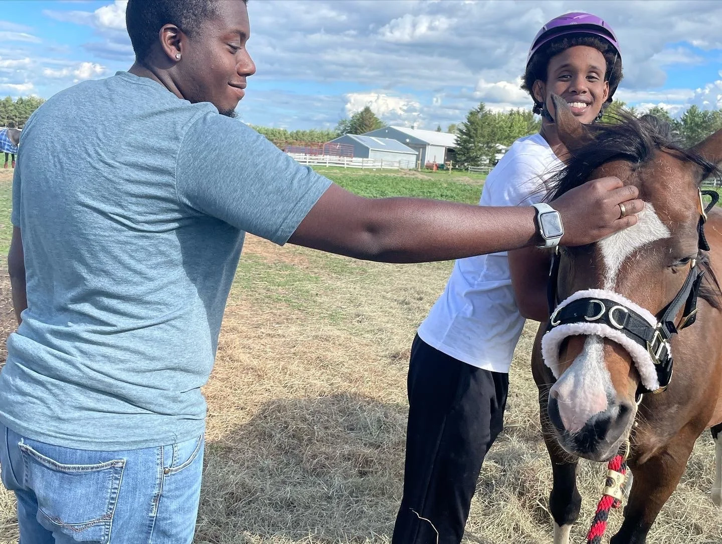 Two people petting a brown horse outdoors with barns and trees in the background.