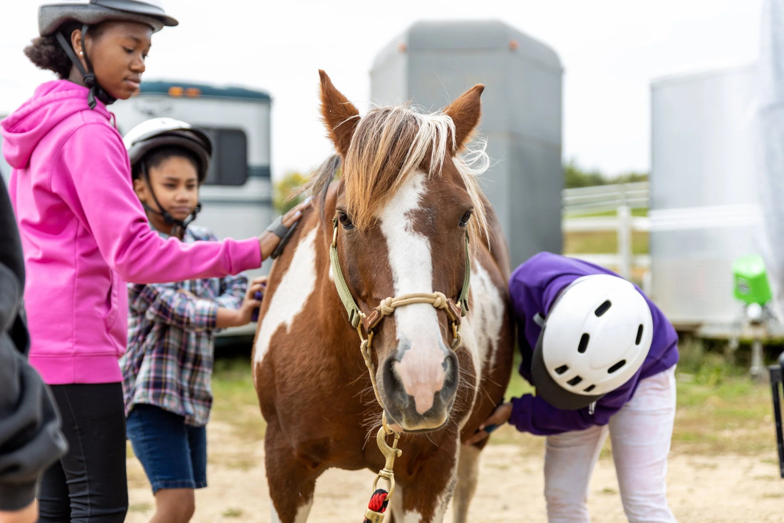 Children wearing helmets standing next to a brown and white horse in an outdoor setting.