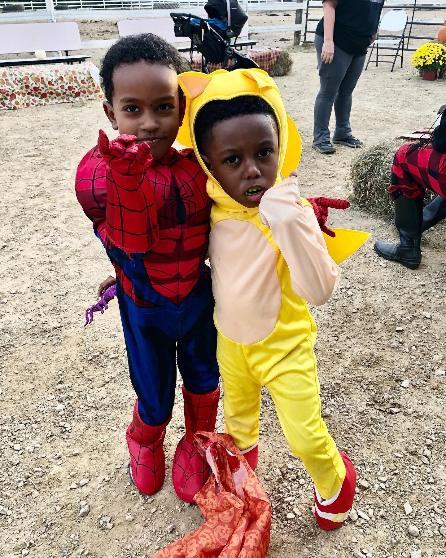 Two children in costumes; one dressed as Spider-Man and the other as Pikachu, posing outdoors on a dirt surface with people and hay bales in the background.