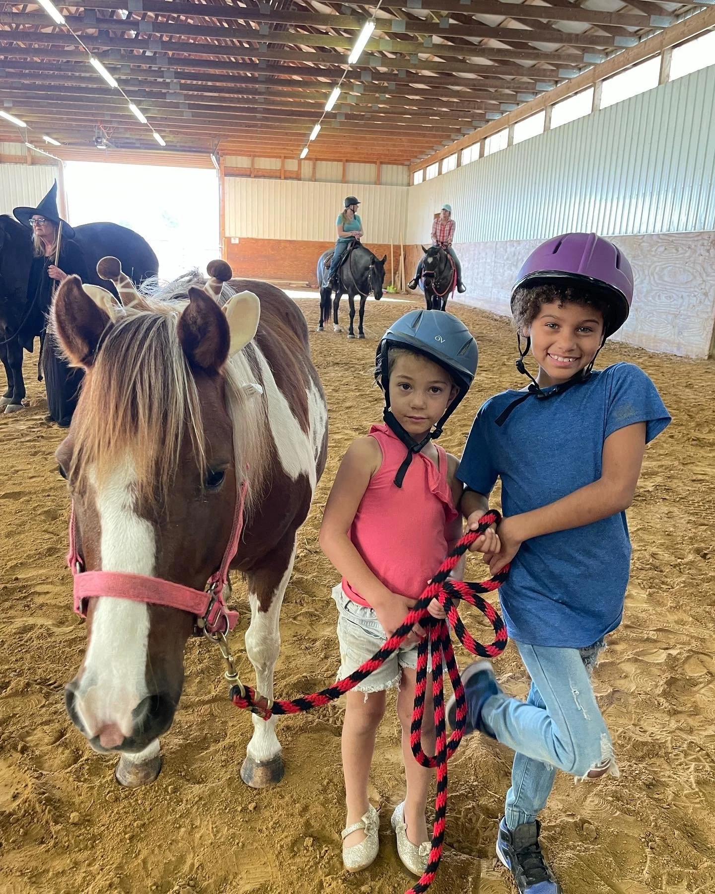 Two children wearing helmets are standing next to a brown and white horse inside an indoor equestrian arena. The children are smiling and holding the horse's lead rope. Several other people and horses can be seen in the background.