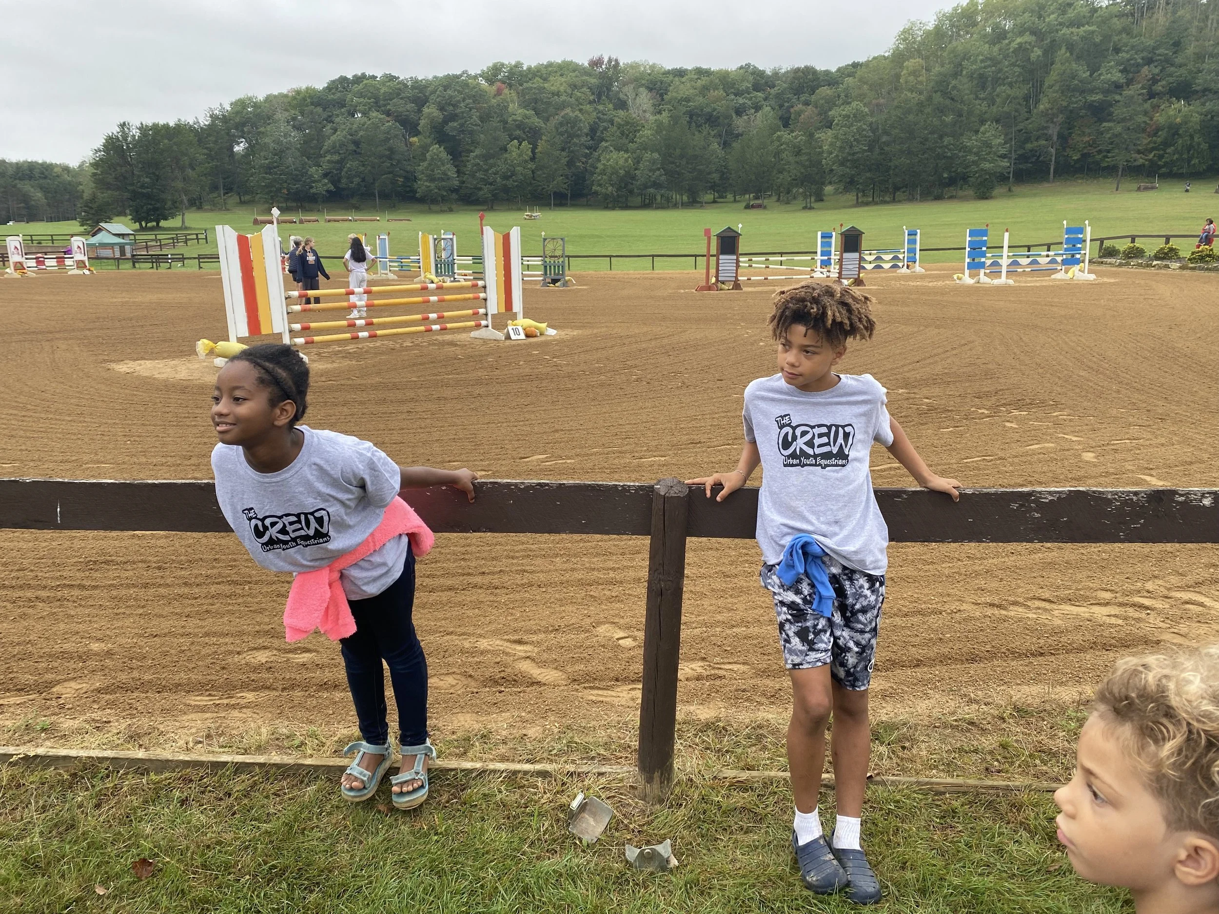 Two children wearing "The Crew - Urban Youth Equestrian" t-shirts stand by a fence at an outdoor equestrian arena, with show jumping obstacles in the background and trees in the distance.