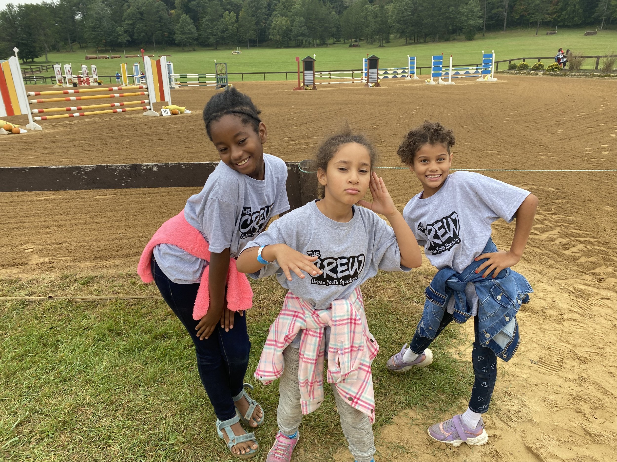 Three children posing with a horse jumping arena in the background. They are wearing matching gray t-shirts, and each has a jacket tied around their waist. The setting seems to be an outdoor equestrian park with various jumping obstacles visible behi