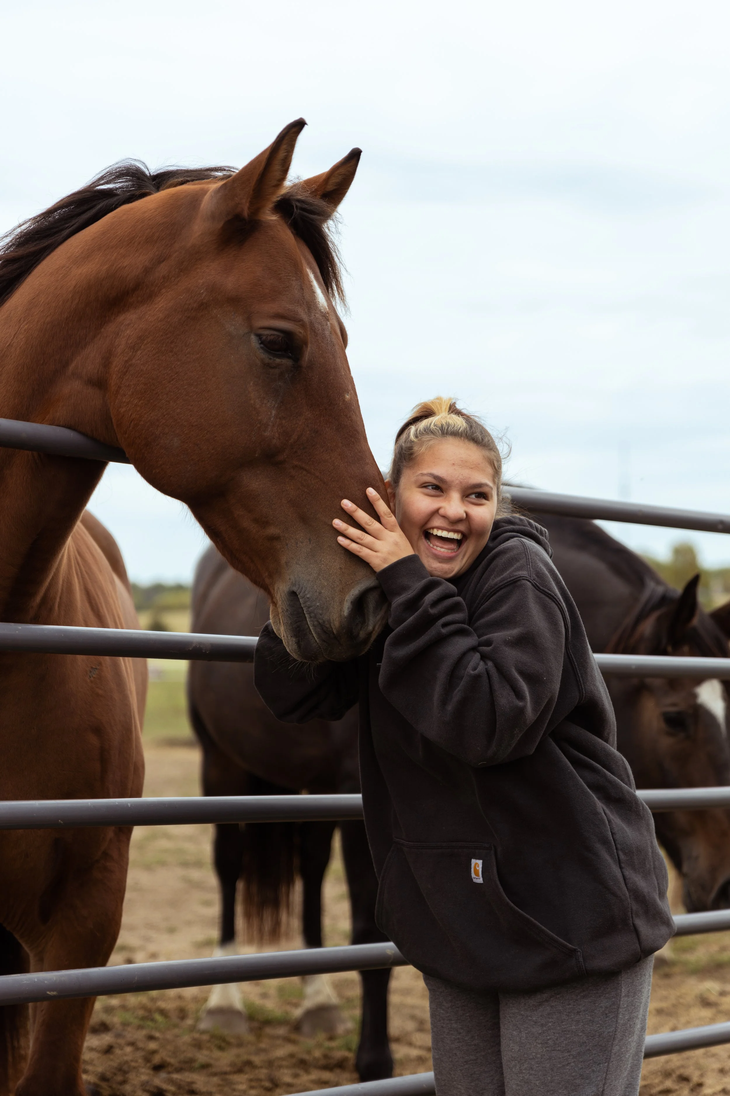 A person smiling and hugging a brown horse near a metal fence, with another horse in the background.