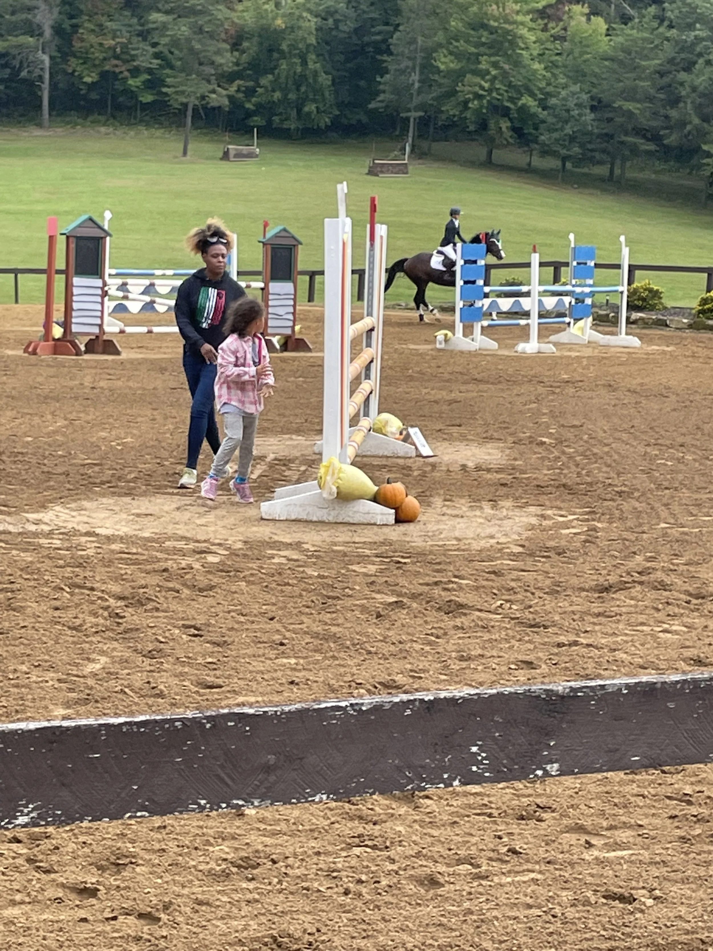 Two people walking on a dirt arena with horse jumps, a rider on a horse in the background, and trees in the distance.