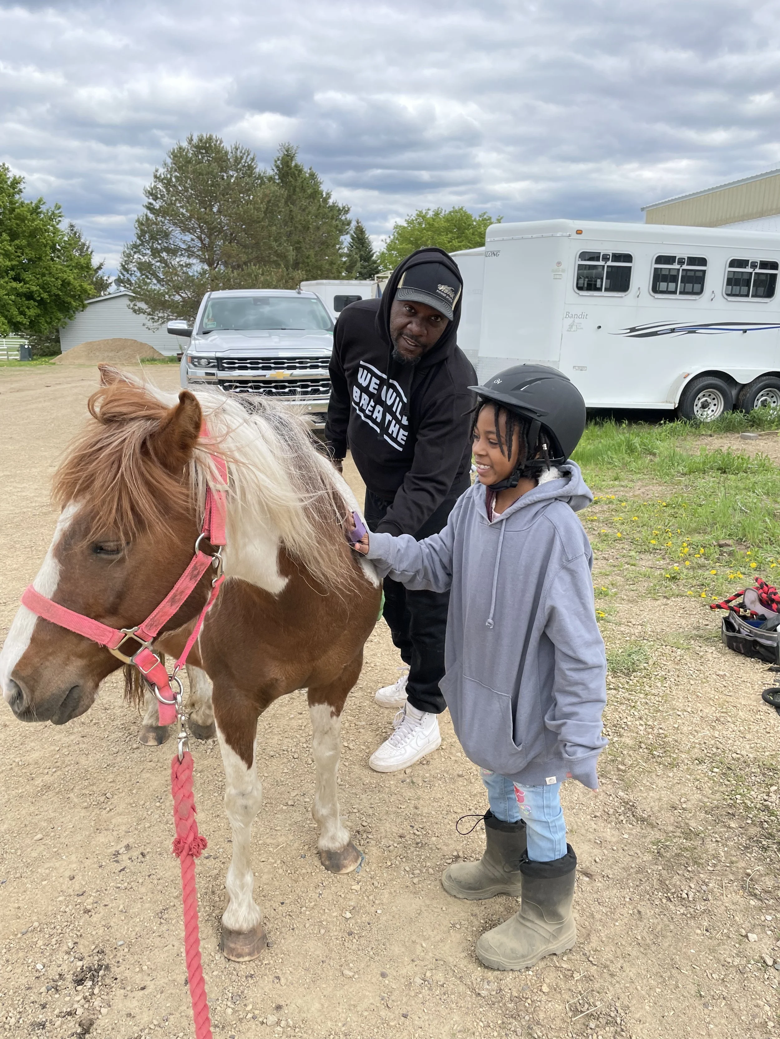 A child wearing a helmet brushing a pony with a man standing nearby, in an outdoor area with a vehicle and a horse trailer in the background.