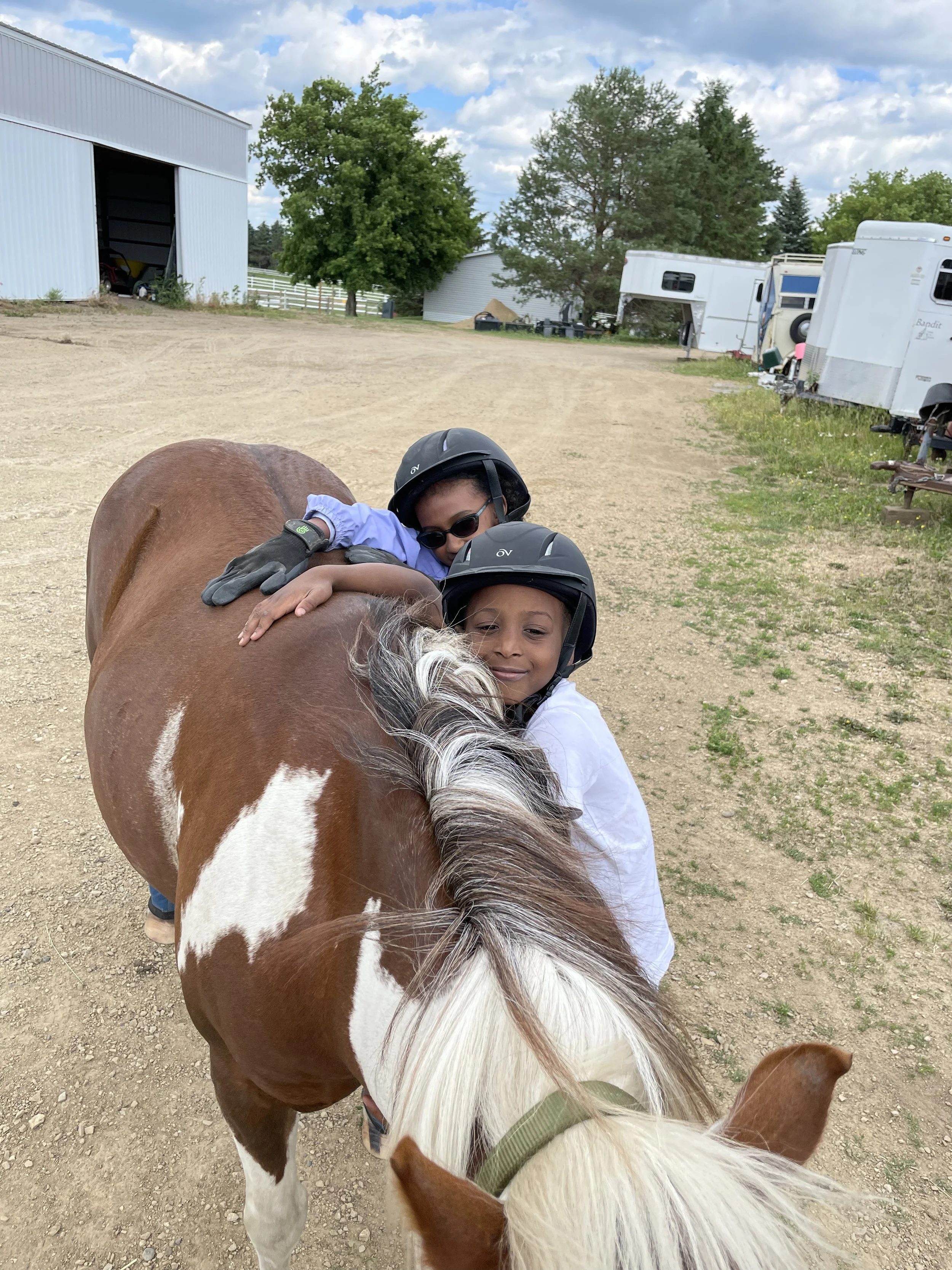 Two children wearing helmets hugging a brown and white horse outside, with trailers, buildings, and trees in the background.