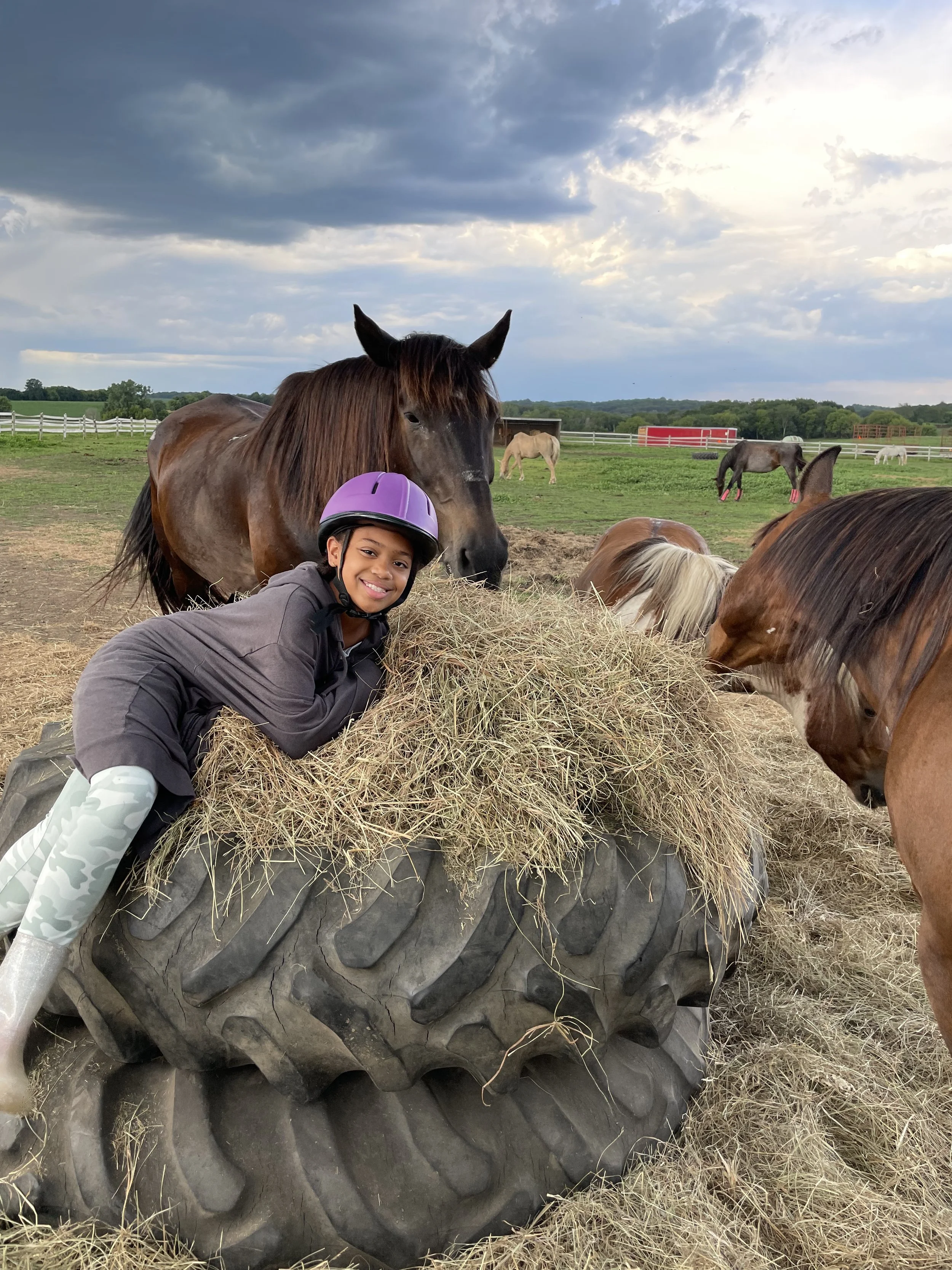 Child wearing a purple helmet lies on hay piled on a large tire with horses in the background in a grassy field under a cloudy sky.