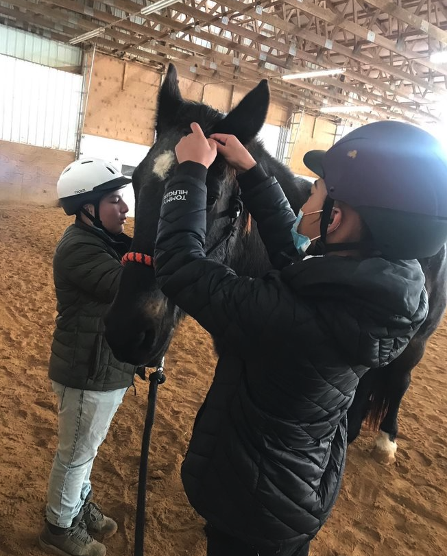 Two people wearing helmets and jackets interacting with a horse inside an indoor riding arena.