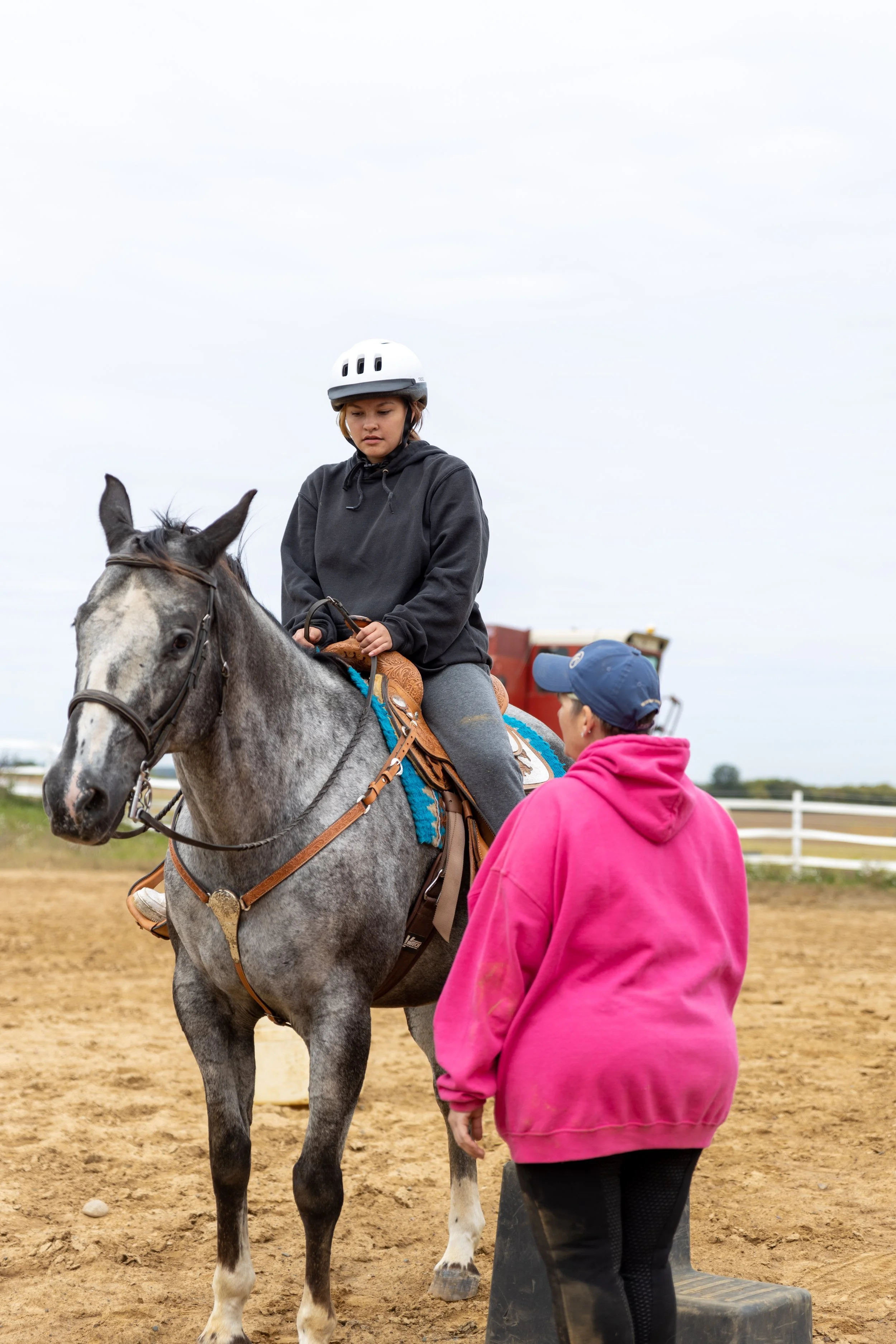 A person wearing a white helmet sits on a gray horse, having a conversation with another person in a pink hoodie at a riding arena.