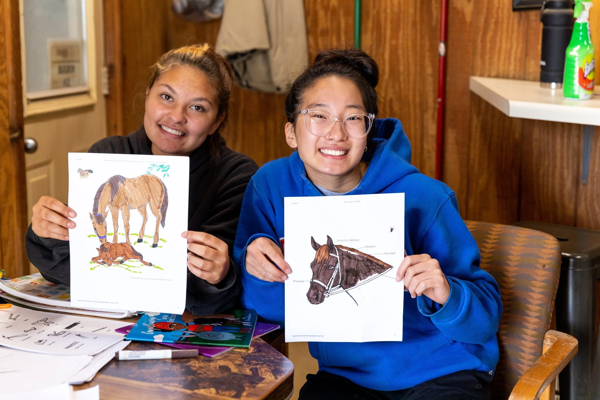 Two people sitting at a table, smiling and showing off colored horse drawings. The person on the left is holding a drawing of a horse and a foal, while the person on the right holds a drawing of a horse's head. They are indoors with a wooden wall and