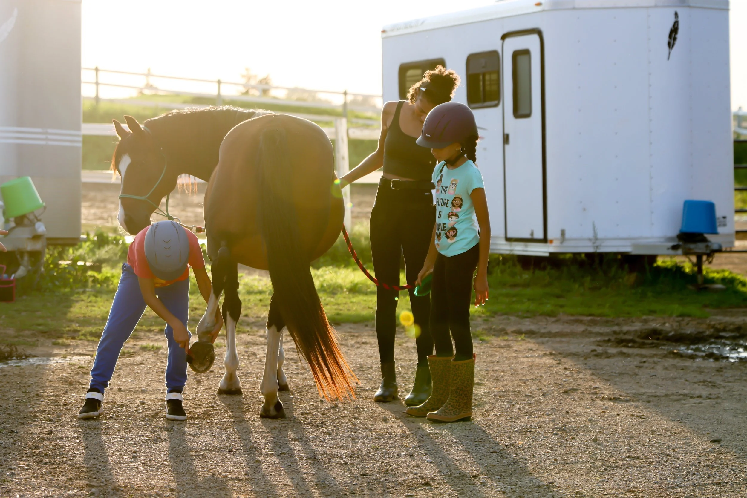 Two children wearing helmets and a woman with a horse, one child cleaning the horse's hoof, near a horse trailer in an outdoor setting.