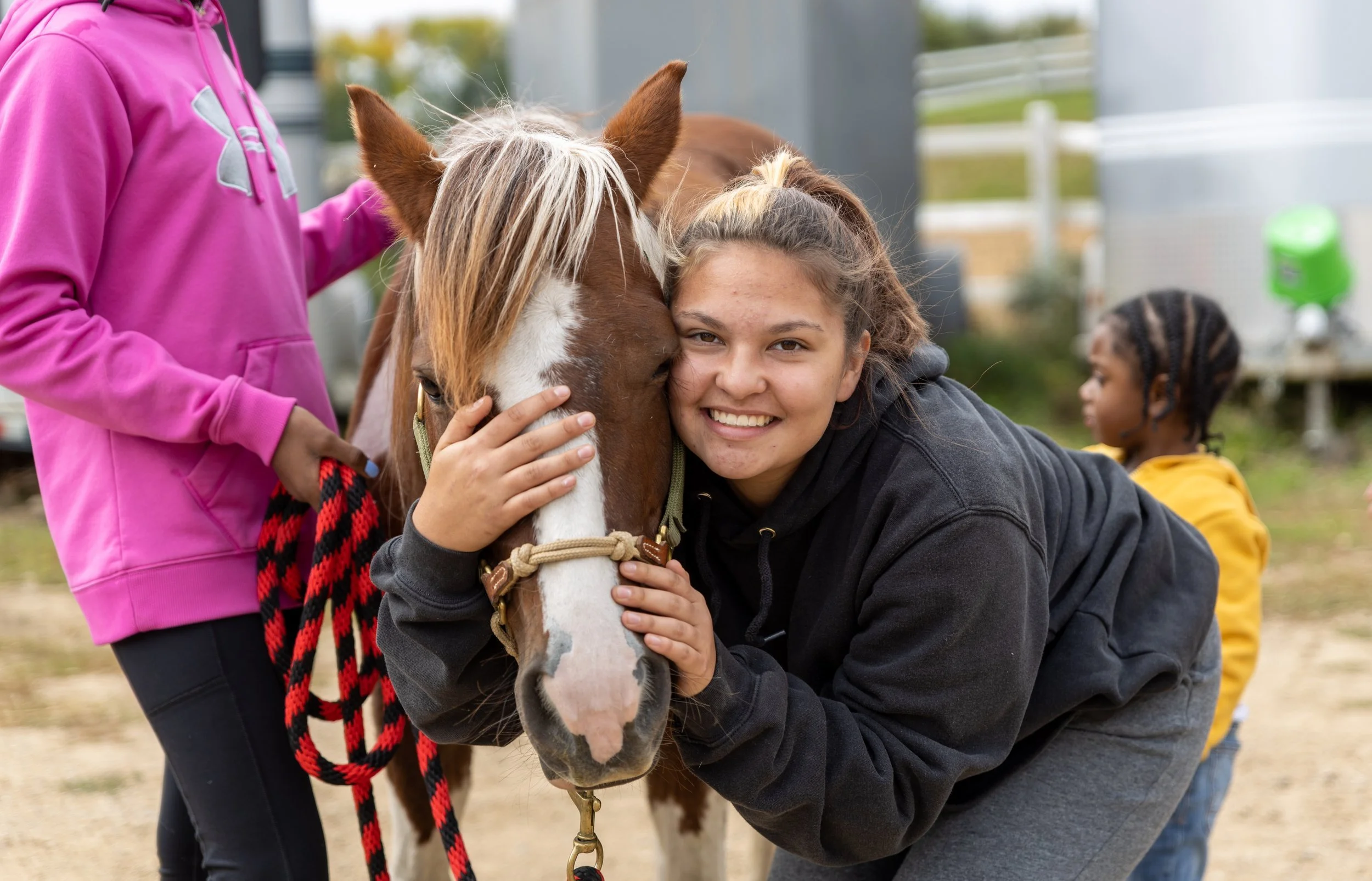 A young woman smiling while hugging a brown and white horse. Another person is holding a rope attached to the horse. A child in a yellow jacket is in the background.