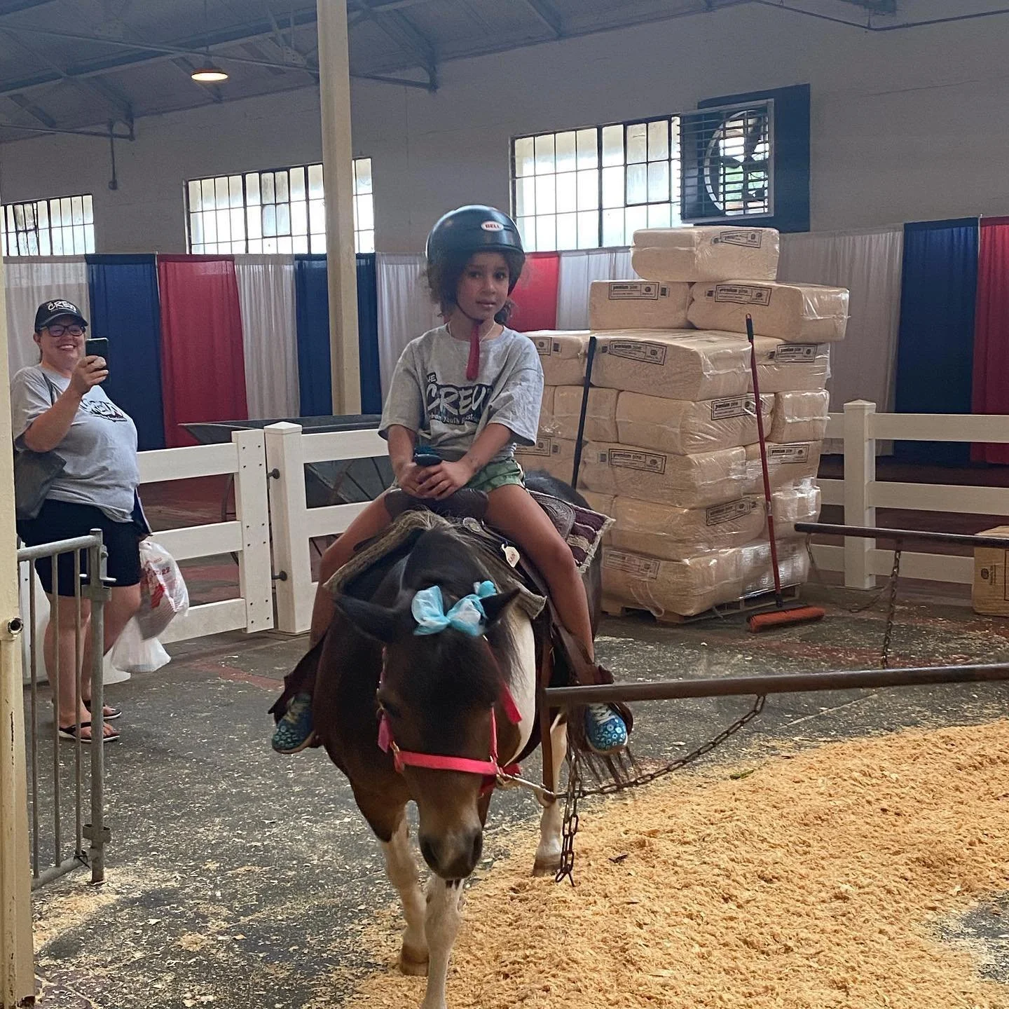 A child riding a pony with a helmet indoors while a person takes a photo. The pony has a blue bow on its head, and there's a stack of packaged hay in the background.