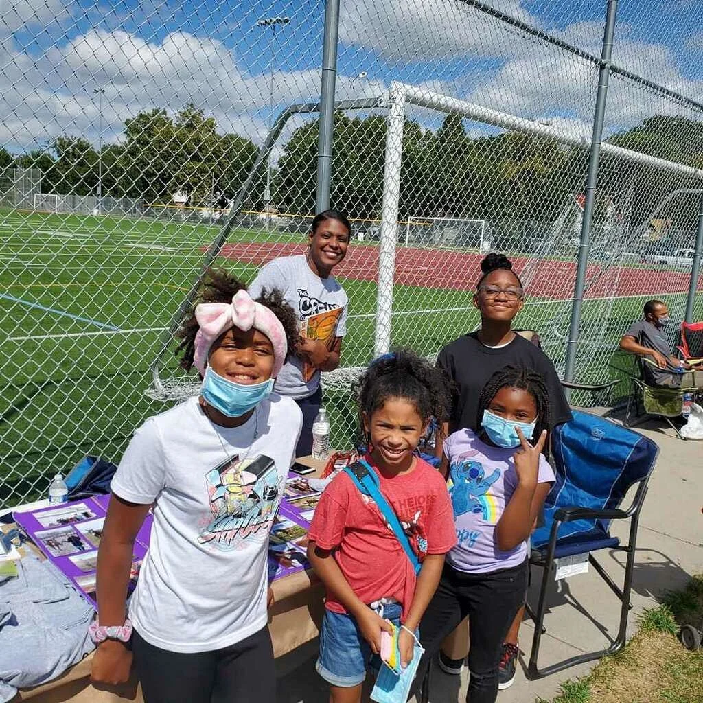 Group of smiling children and an adult standing by a table at a sports field, with a chain-link fence and grassy area in the background.