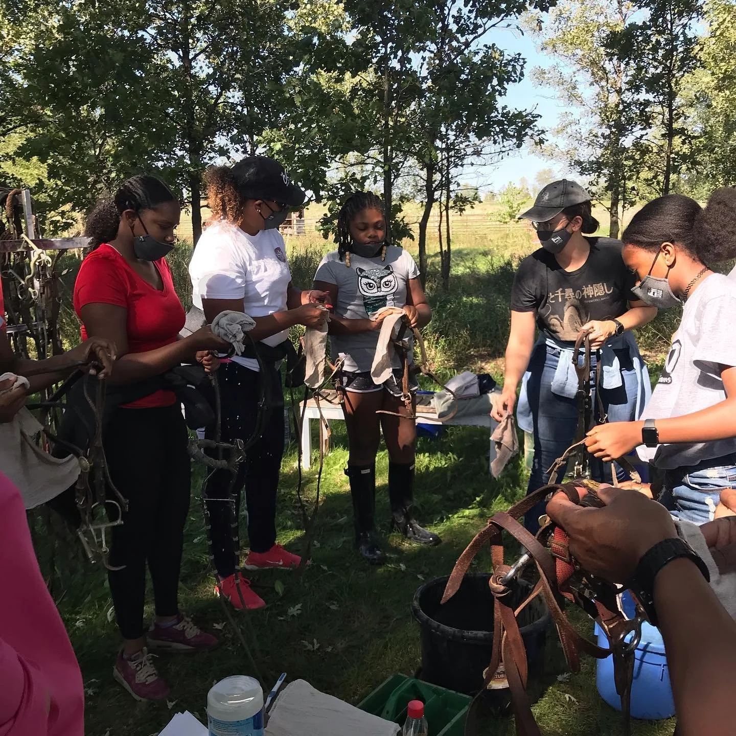 Group of people outdoors cleaning horse bridles, wearing masks and casual attire, surrounded by trees and greenery.