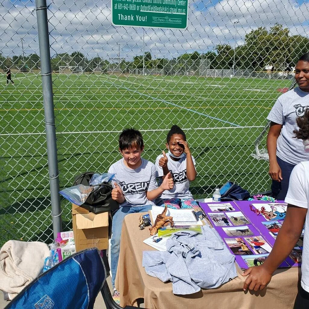 Children sitting behind a table with crafts and photos, smiling and giving thumbs up, near a fenced sports field.