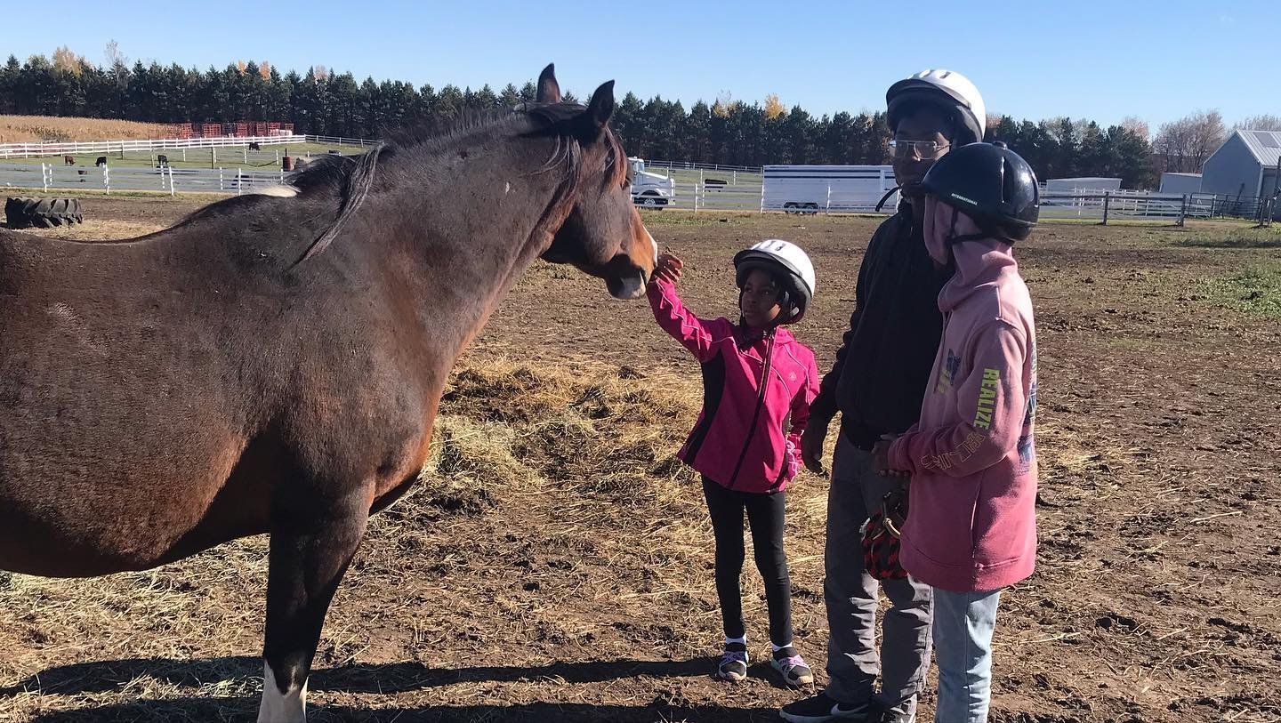 Three people, including a child, wearing helmets petting a brown horse in a farm setting.