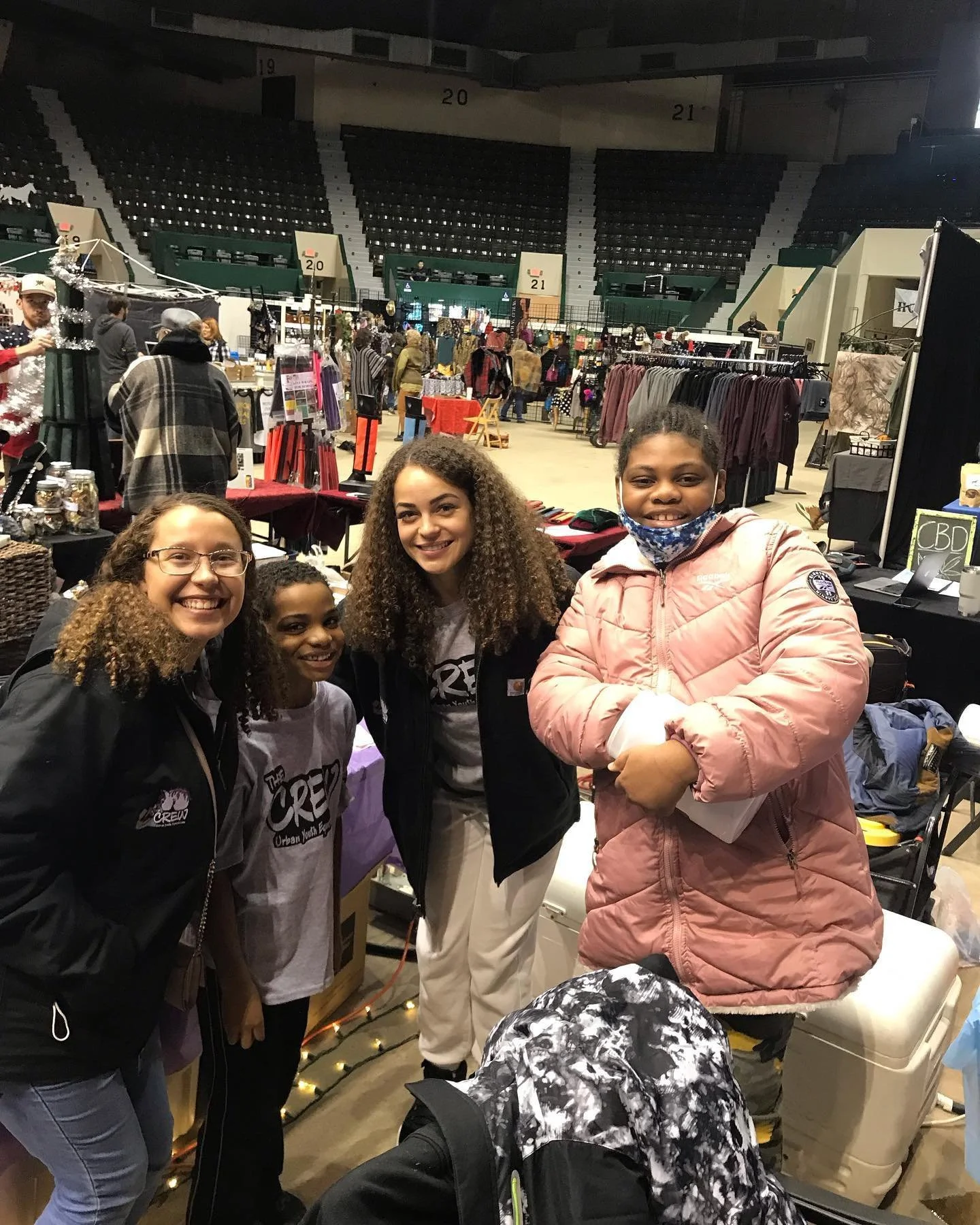 Group of people smiling at a market event with various stalls selling clothing and other items in the background.