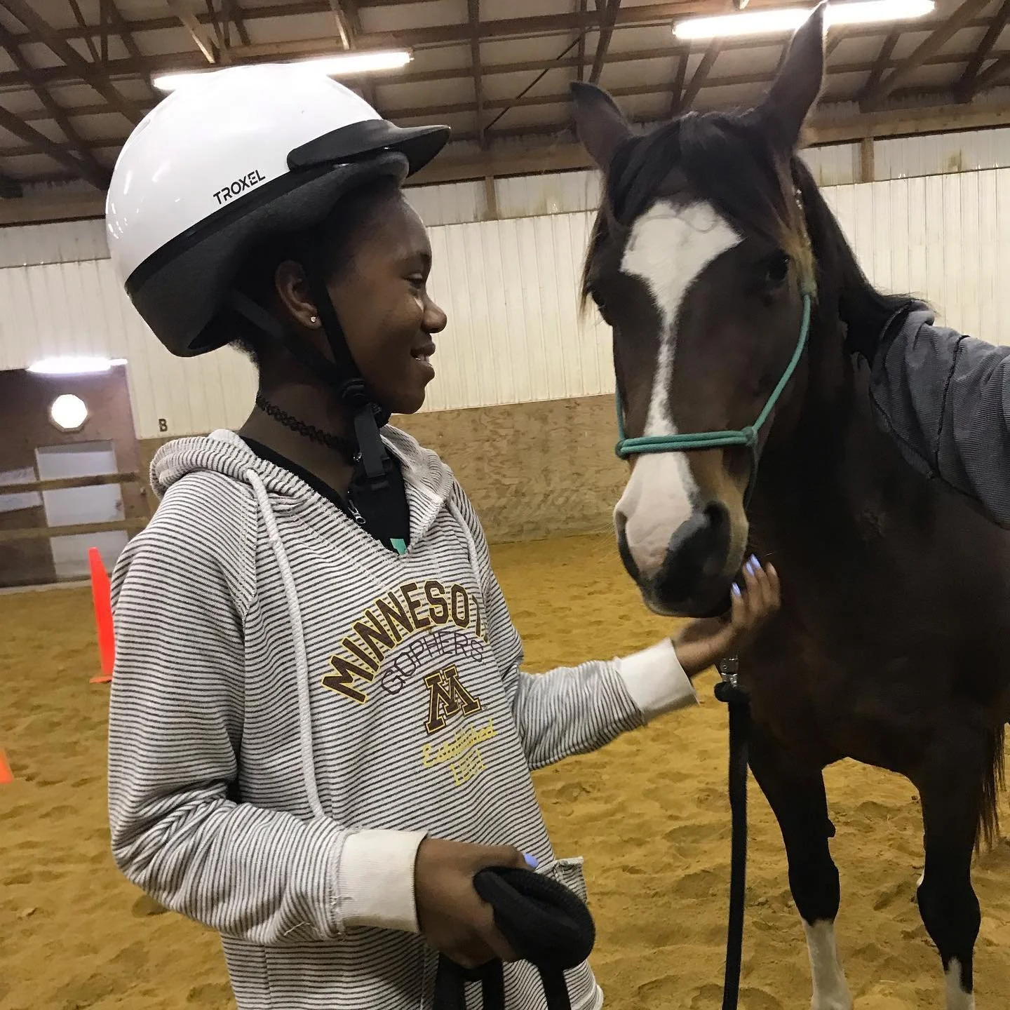 Person wearing a helmet petting a brown horse in an indoor arena.