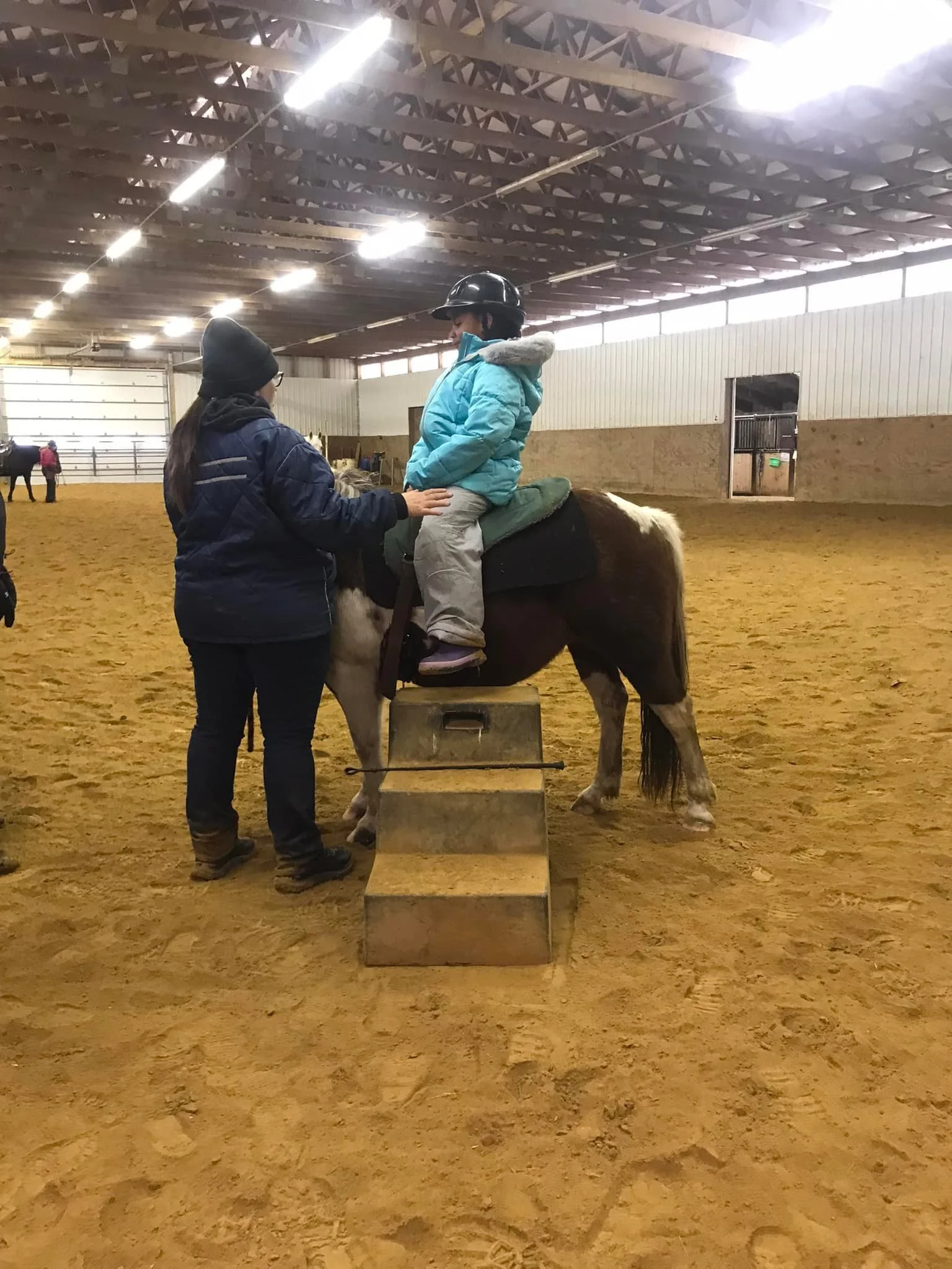 Child in a blue jacket and helmet sitting on a pony next to an instructor in an indoor riding arena.