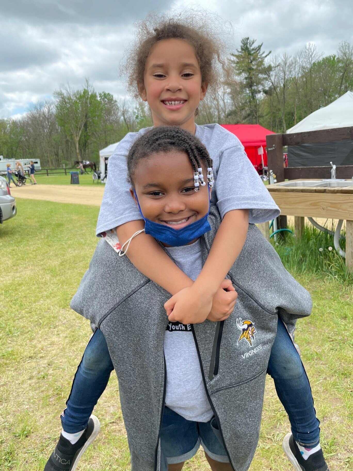 Two children outdoors, one giving a piggyback ride, surrounded by grass and tents.