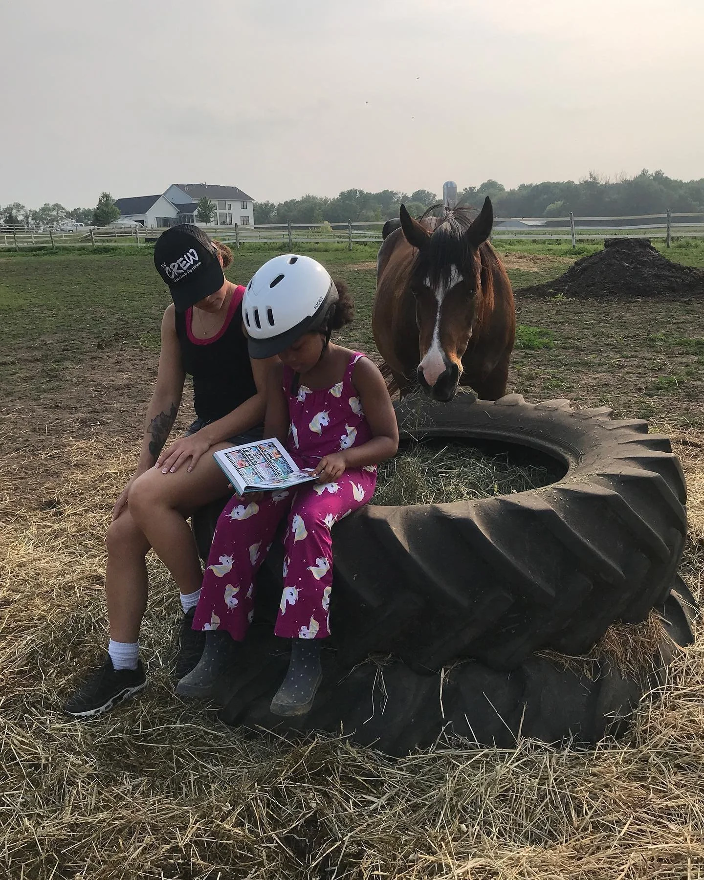 A woman and a child sitting on a large tractor tire reading a book outdoors. The child is wearing a safety helmet. A horse is standing next to them in a grassy field. A farmhouse and trees are visible in the background.