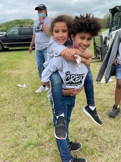 Two children wearing matching "The Crew" shirts having fun outdoors, with one child giving the other a piggyback ride on a grassy field. Part of a group, possibly at a community event.