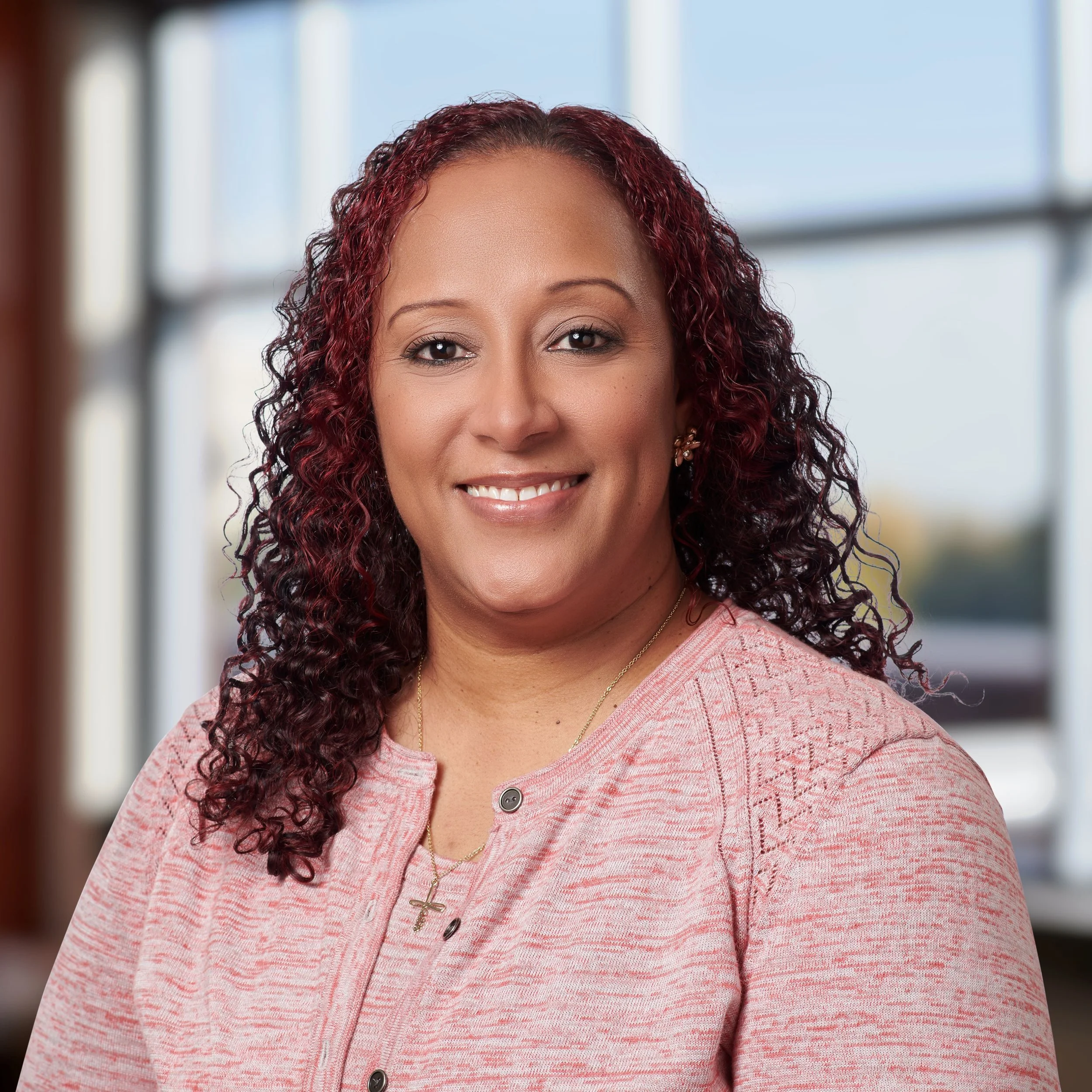 Smiling person with curly hair wearing a pink top, indoors with a window background.