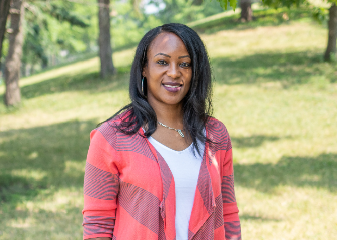 Woman in outdoors wearing orange striped sweater and white shirt, with trees in the background.