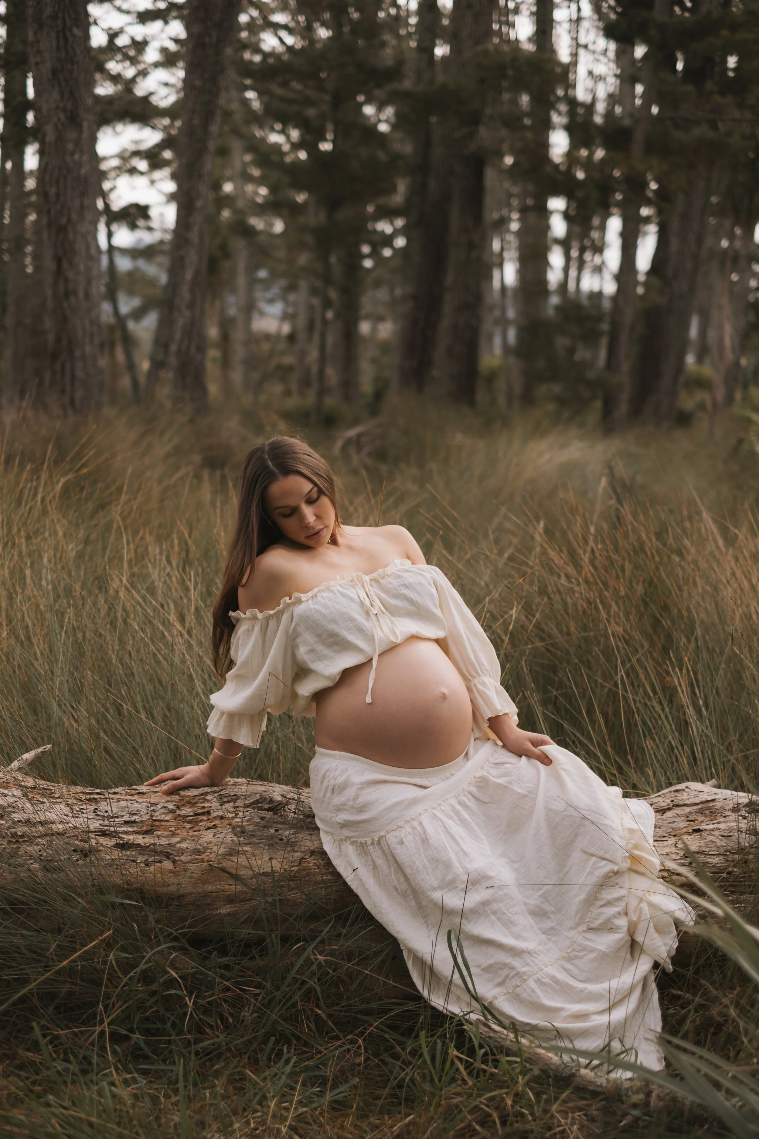 A pregnant woman in a white off-the-shoulder dress sitting on a log in a grassy field with trees in the background, looking down and gently touching her baby bump.