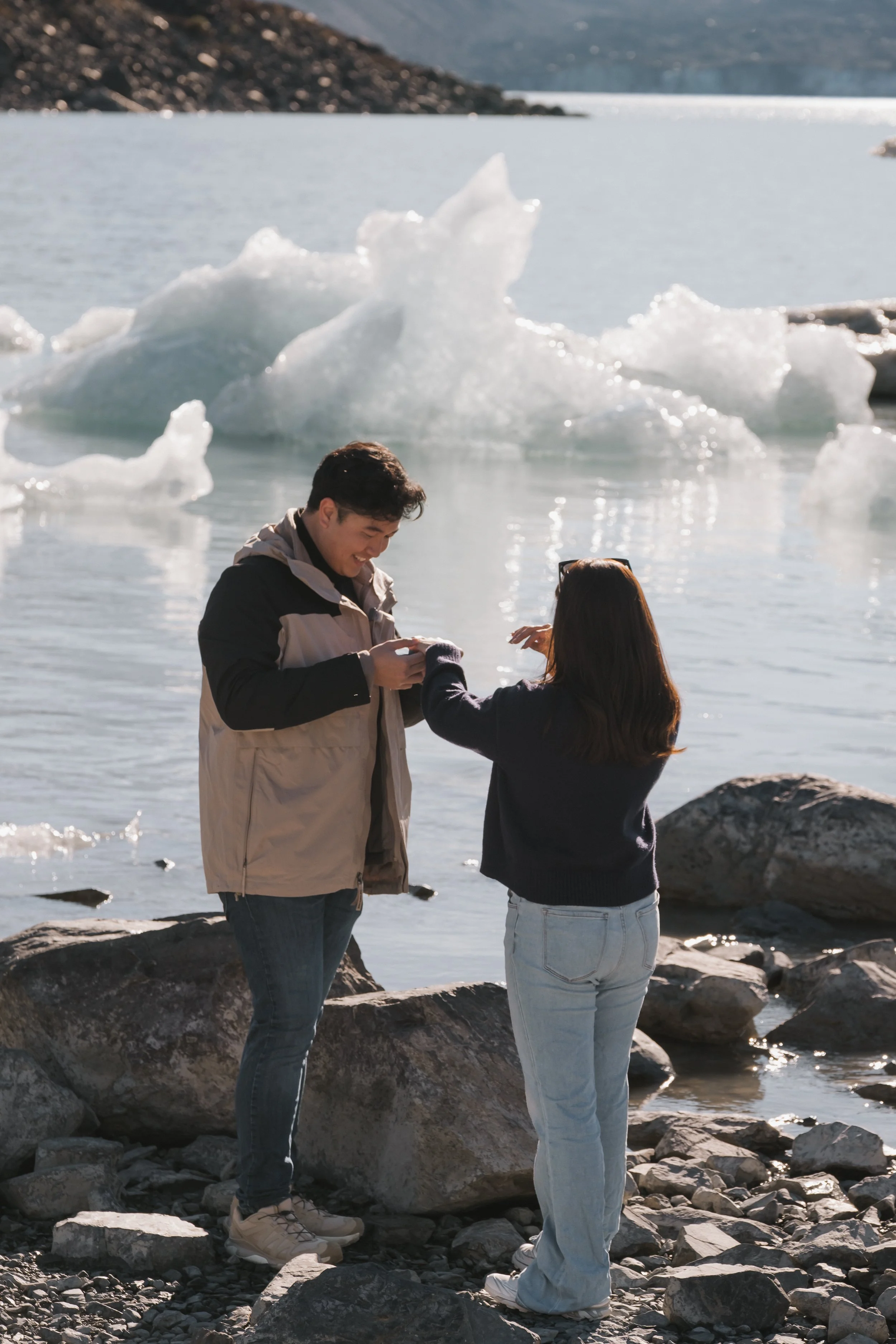 A man and woman standing on rocks by a body of water with floating ice in the background, smiling and interacting during daylight.