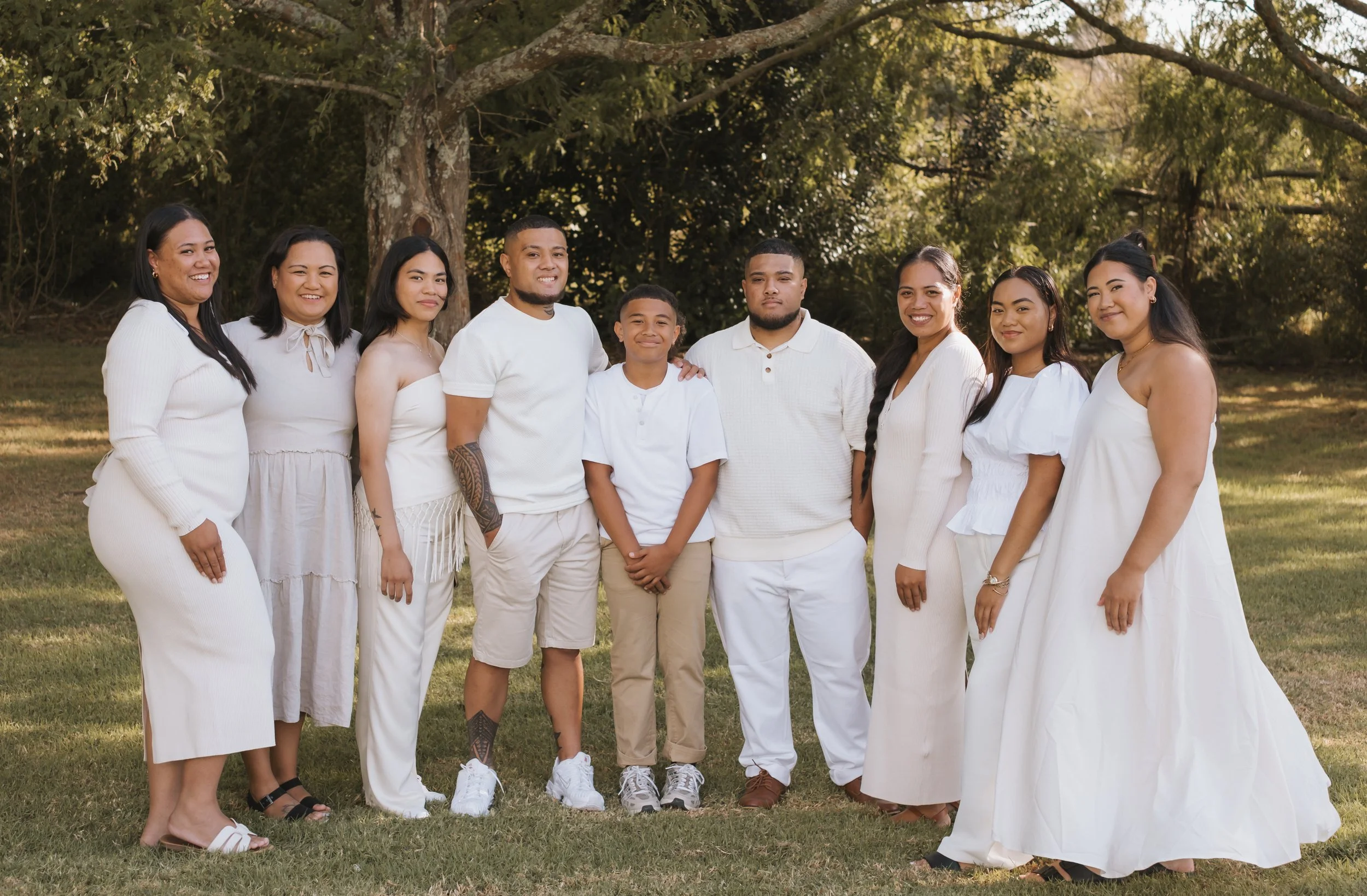 Group of ten people standing outdoors on grass in front of trees, dressed in white clothing, smiling and posing for the photo.