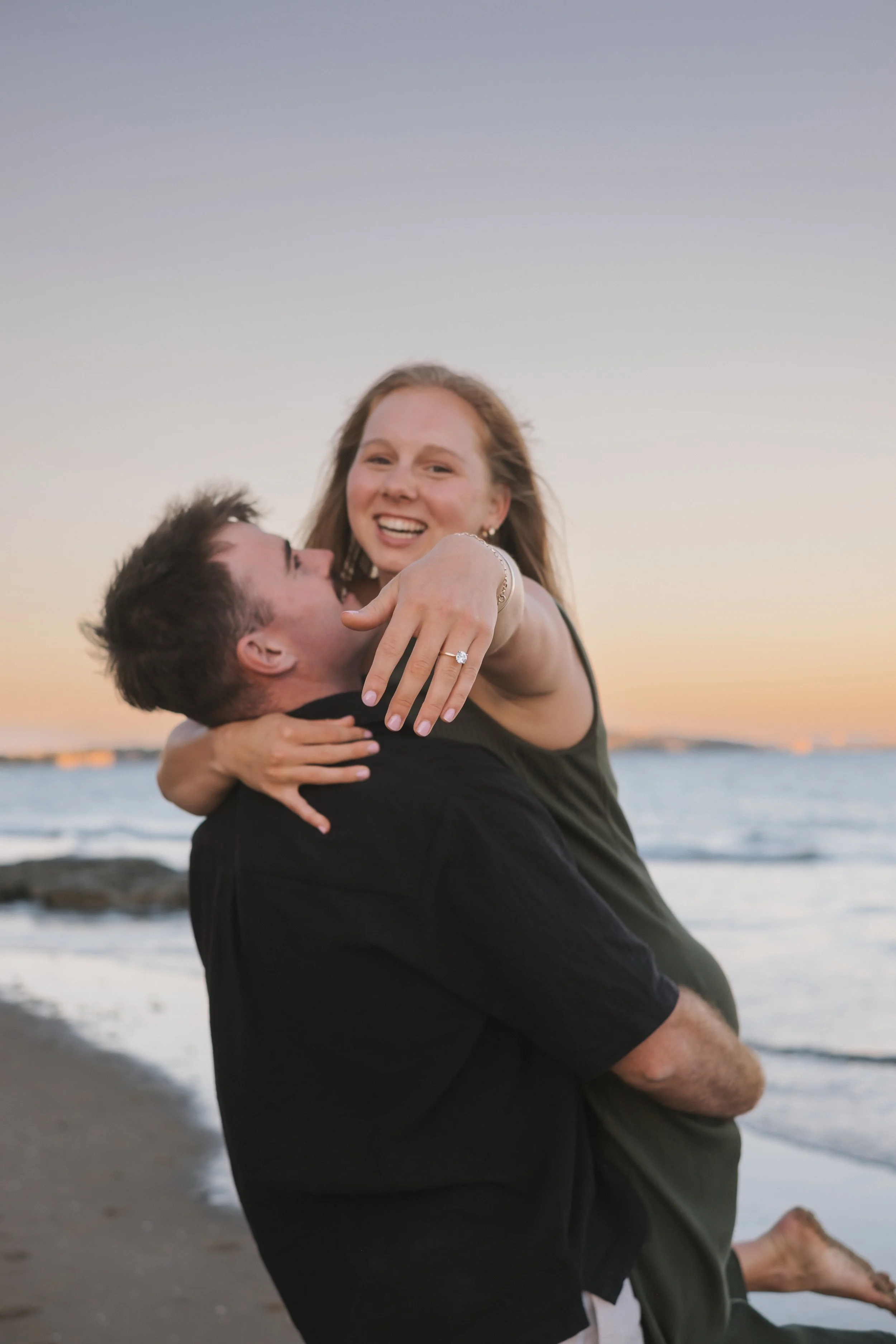 A happy couple at the beach during sunset, with the woman showing off an engagement ring on her finger.