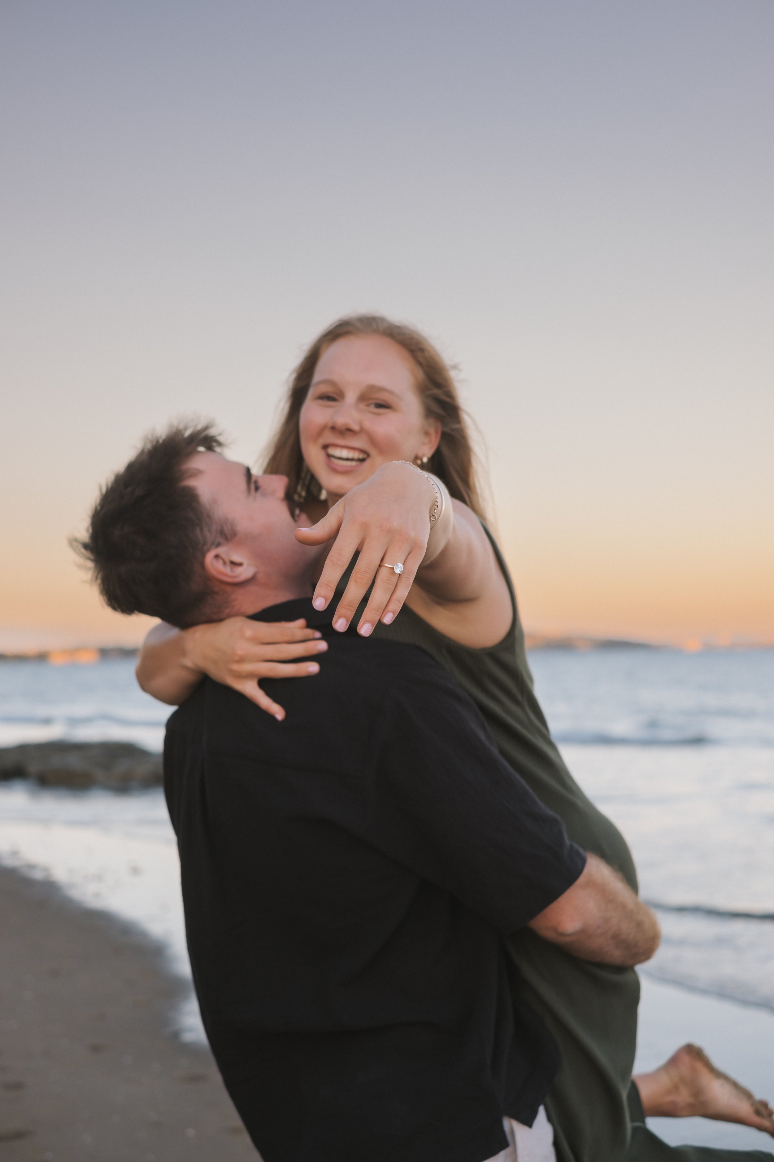 A happy couple at the beach during sunset, with the woman showing off an engagement ring on her finger.