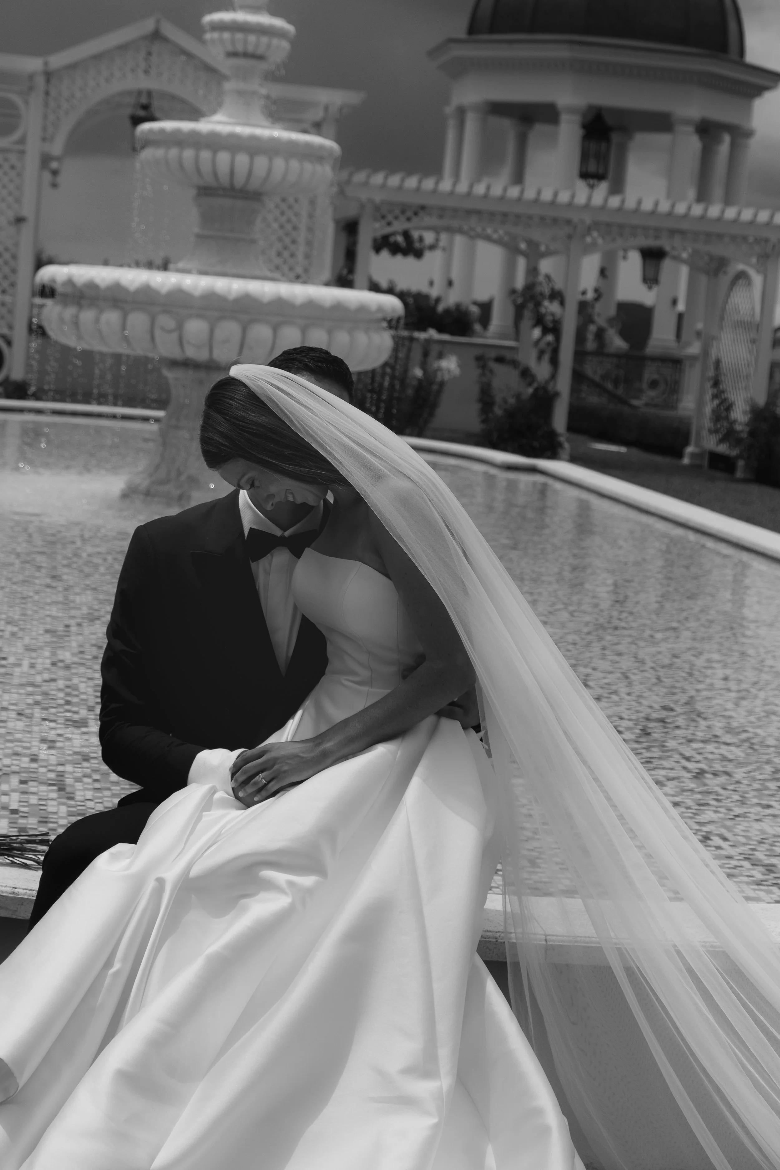 A bride and groom sharing a moment outdoors in front of a large fountain, with the bride wearing a wedding dress and veil, and the groom dressed in a tuxedo.