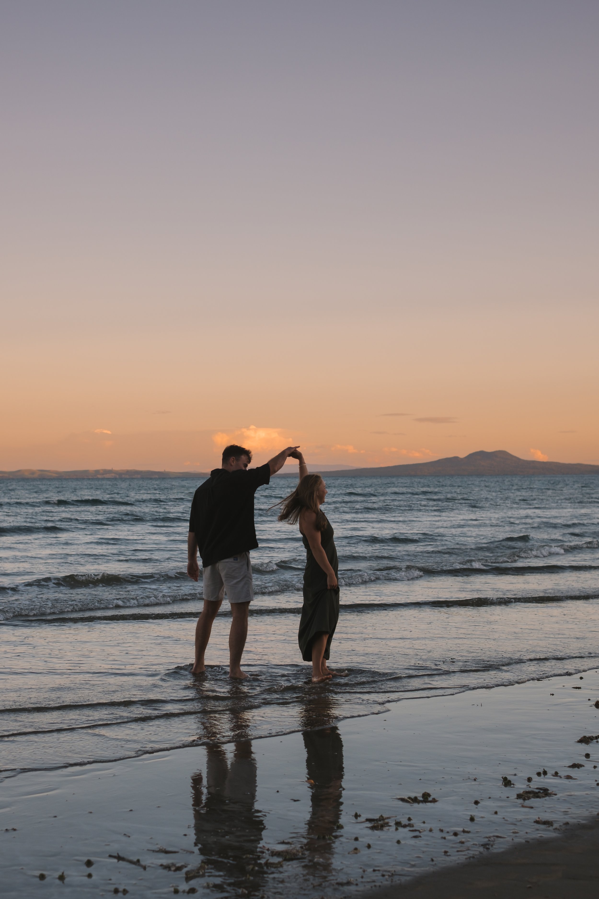 A couple stands in the shallow water at the beach during sunset. The man is holding the woman's hand, and both are facing each other with their feet in the water. The sky is pastel-colored, and mountains can be seen in the distance.