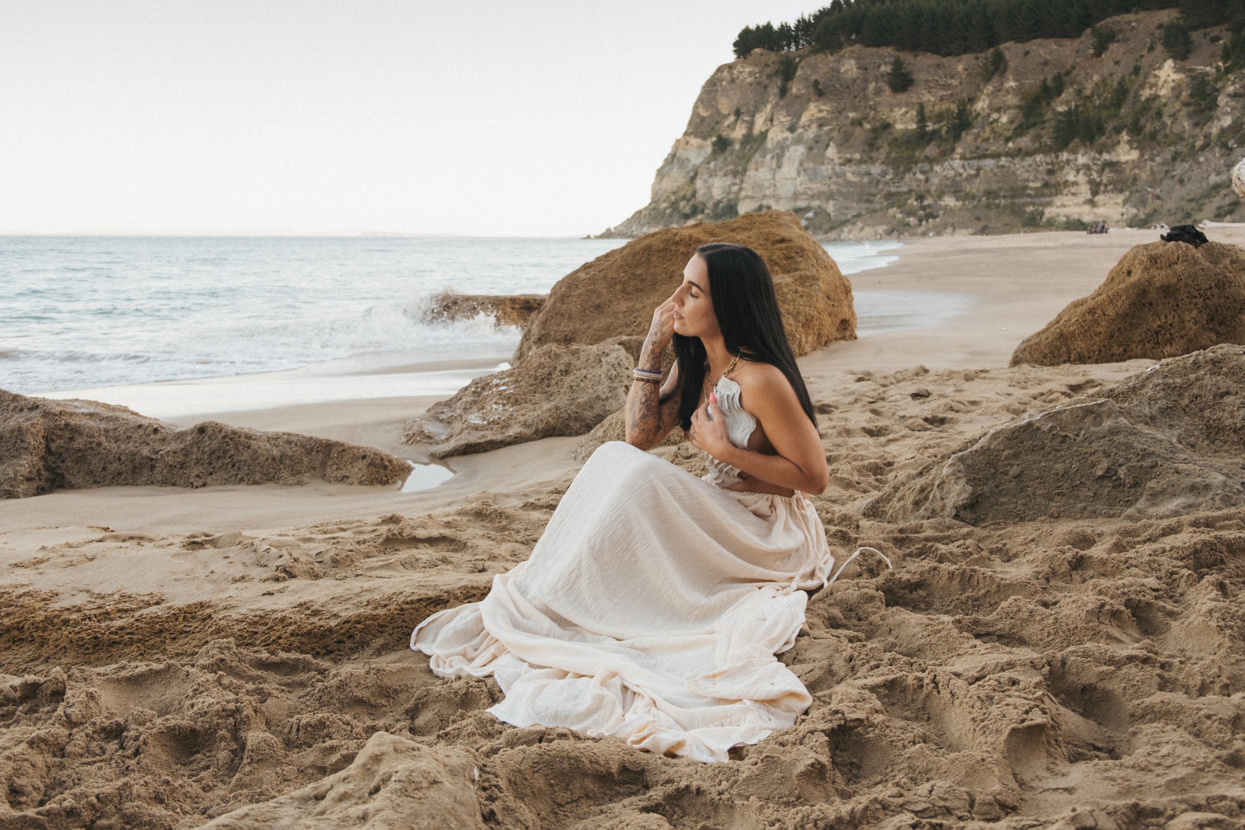Woman sitting on a sandy beach with rocky cliffs, wearing a light dress, near the ocean.