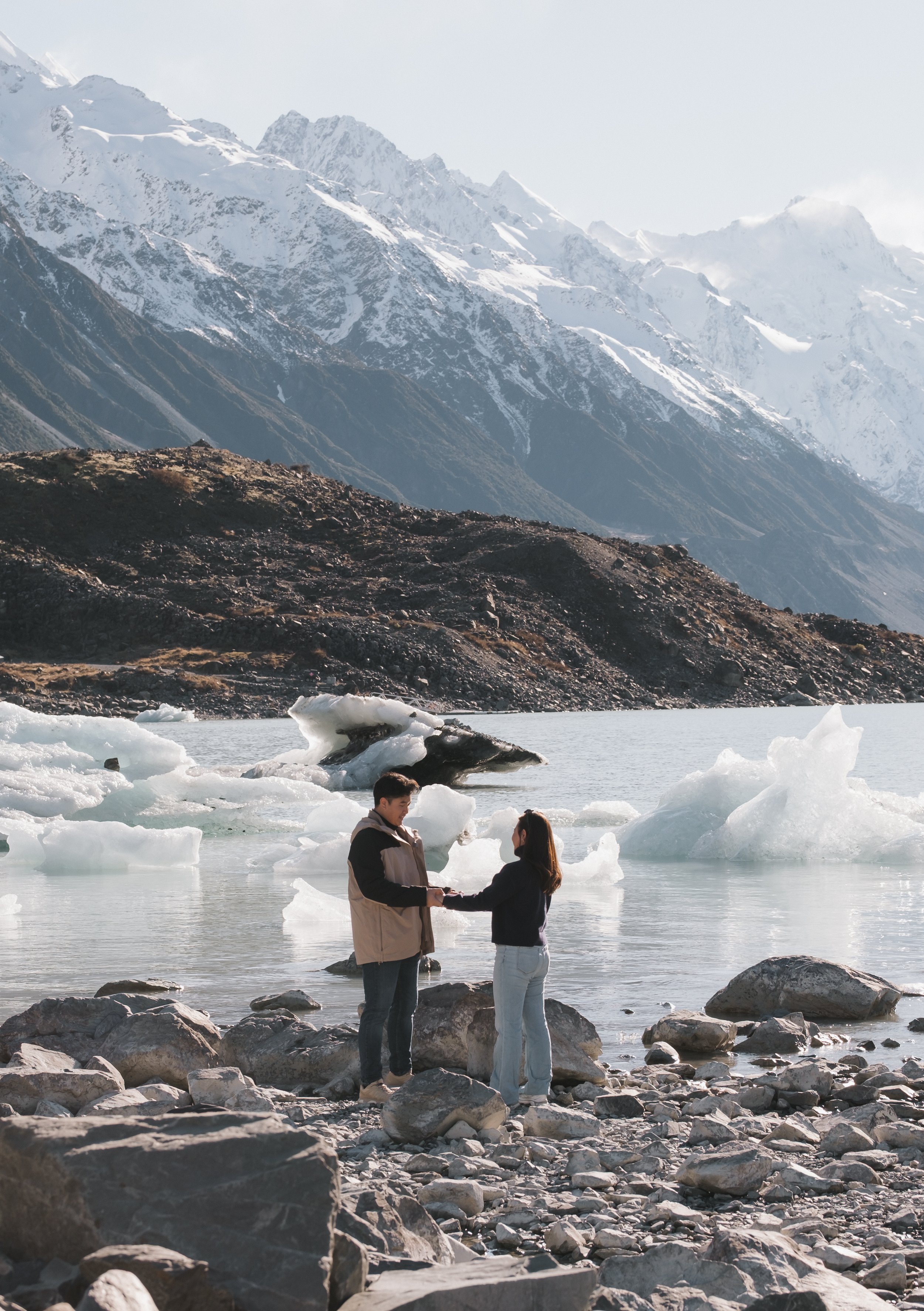 A couple standing on rocks by a lake with icebergs, holding hands and facing each other, with snow-covered mountains in the background.