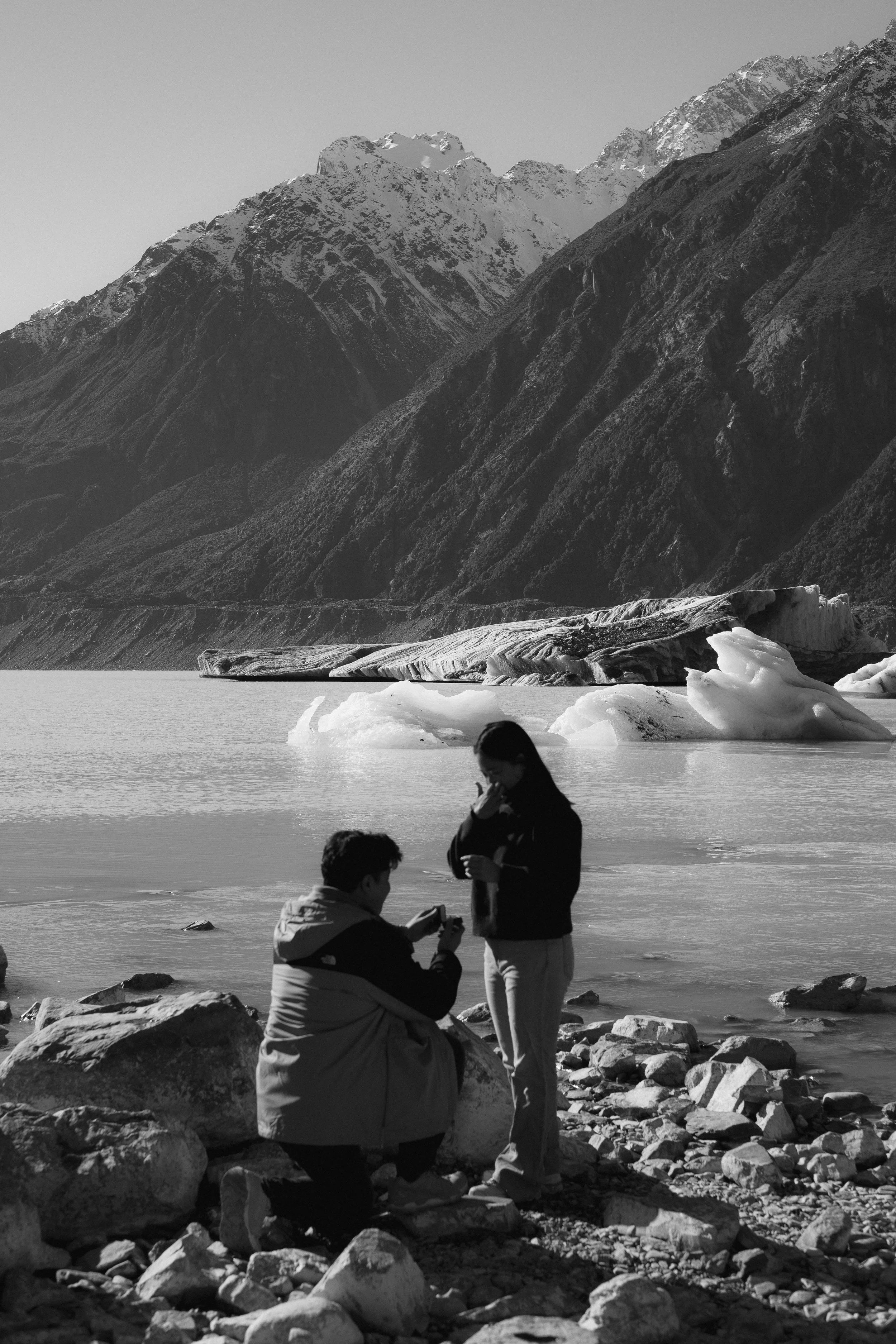 Three people by a rocky lake with snow-capped mountains in the background, in black and white.
