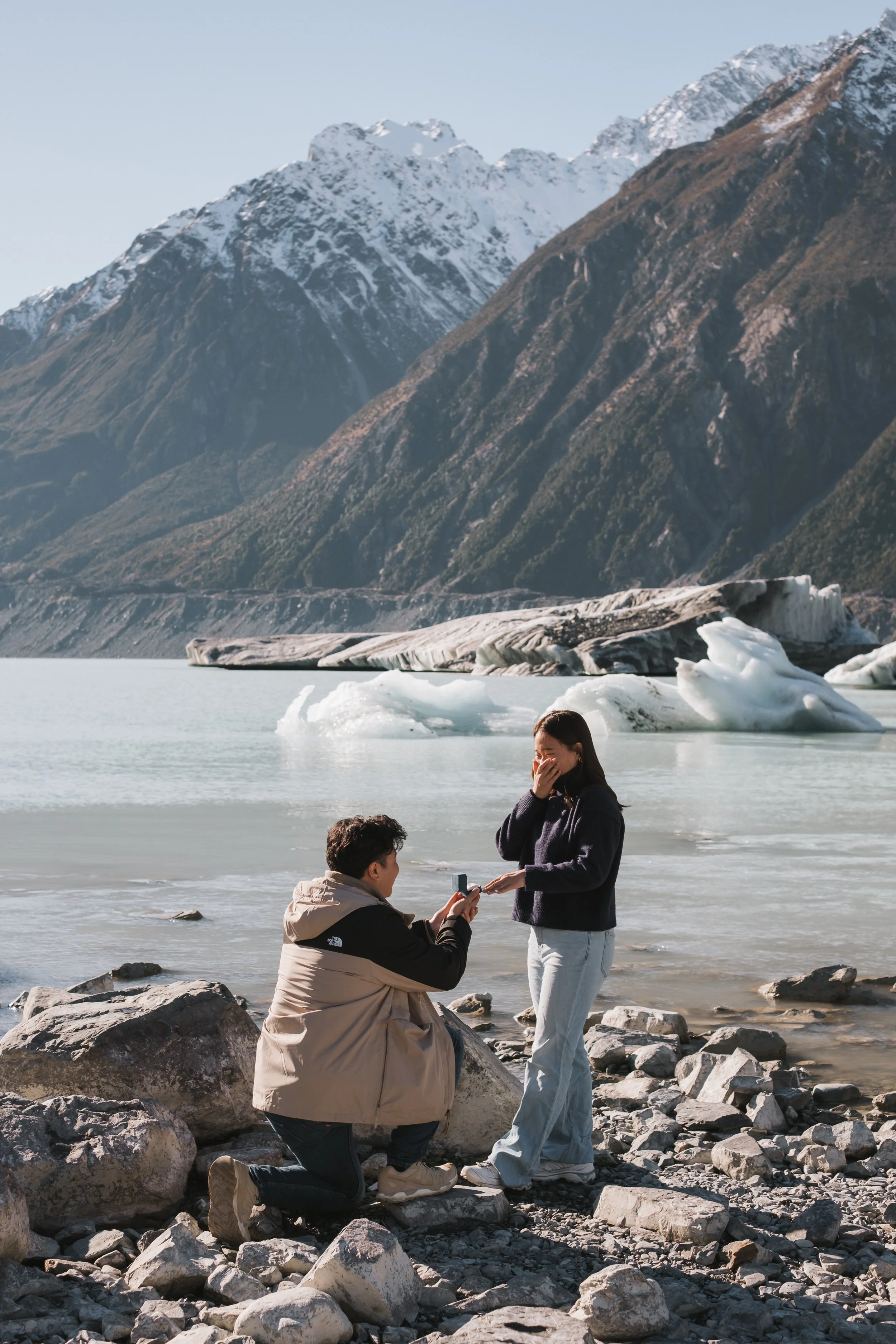 A man in a beige North Face jacket and kneeling on rocks while proposing to a woman in light blue jeans and a dark sweater, with snow-capped mountains and icebergs in the background.