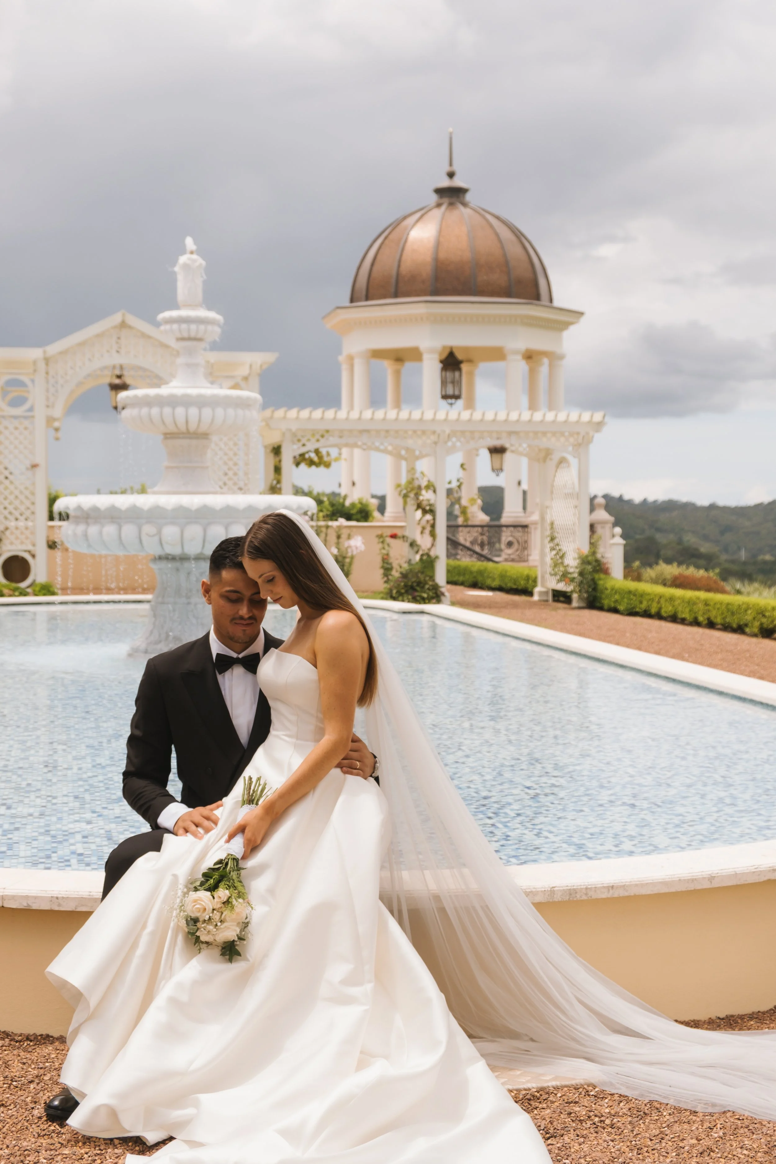 A bride and groom pose together in front of a fountain and a white architectural backdrop, with the bride holding a bouquet of white roses and the groom wearing a black tuxedo with a bow tie.