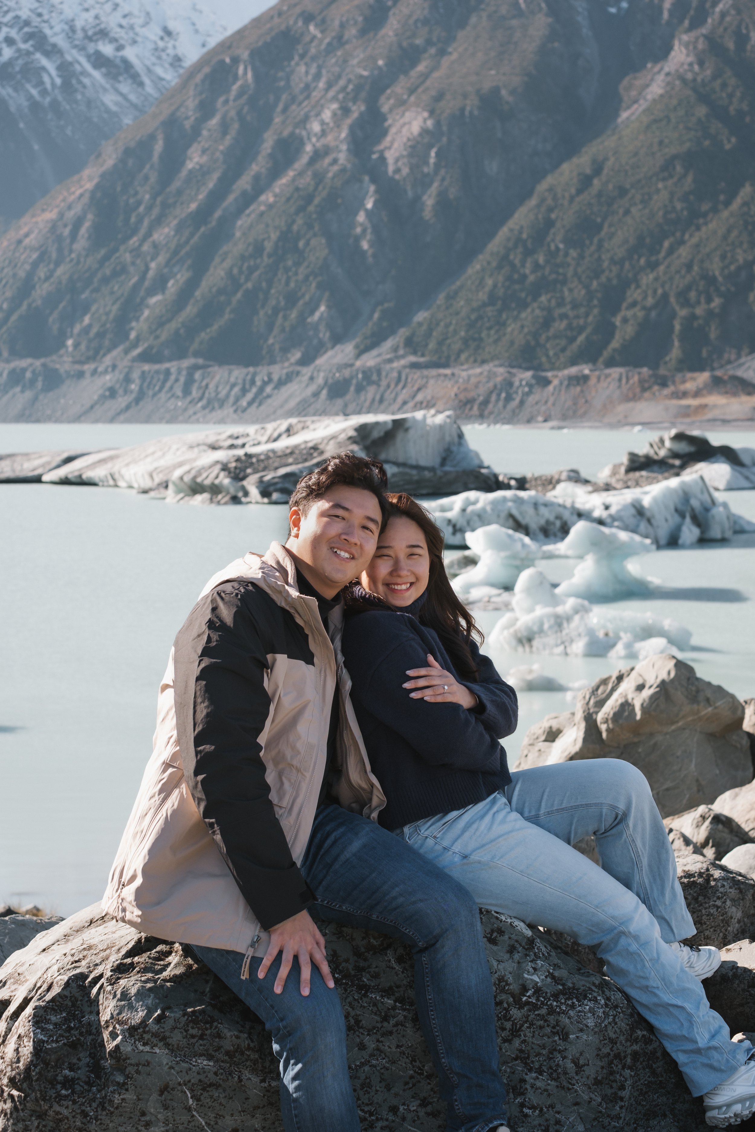 A young man and woman sitting on rocks by a glacier lake, with snow-capped mountains in the background, smiling and enjoying outdoor scenery.