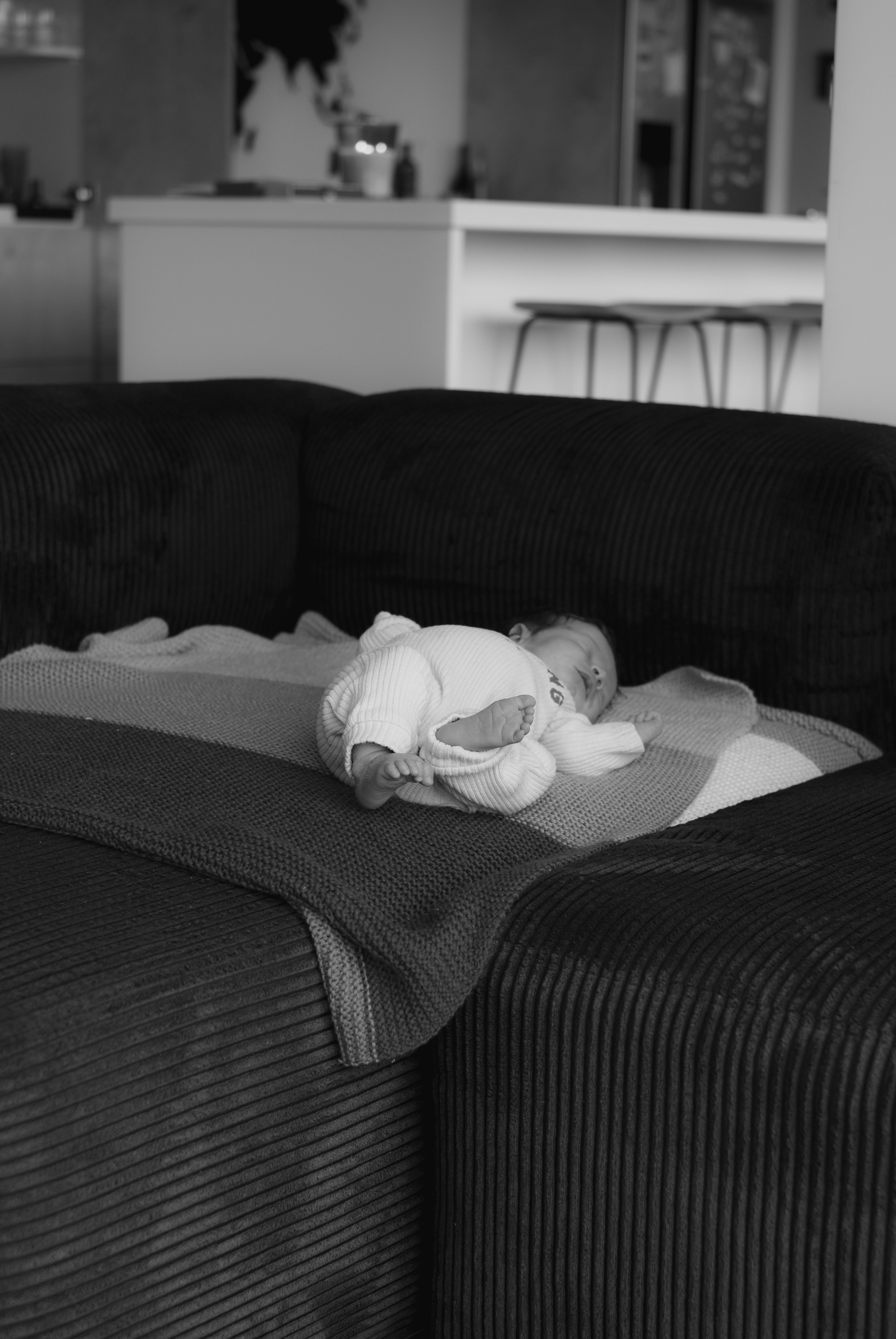 A black and white photo of a baby sleeping on a couch with a blanket, under a dark, textured blanket.