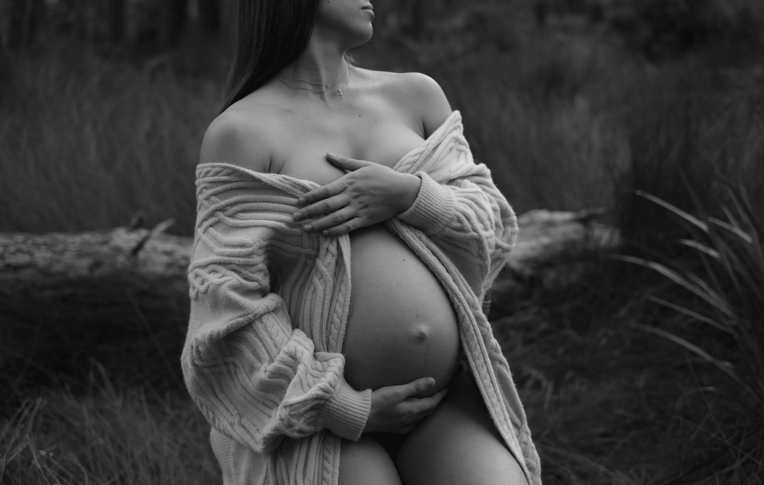 A black and white photo of a woman outdoors, partially covered with a cozy striped sweater, holding her pregnant belly with one hand and crossing her arms over her chest.