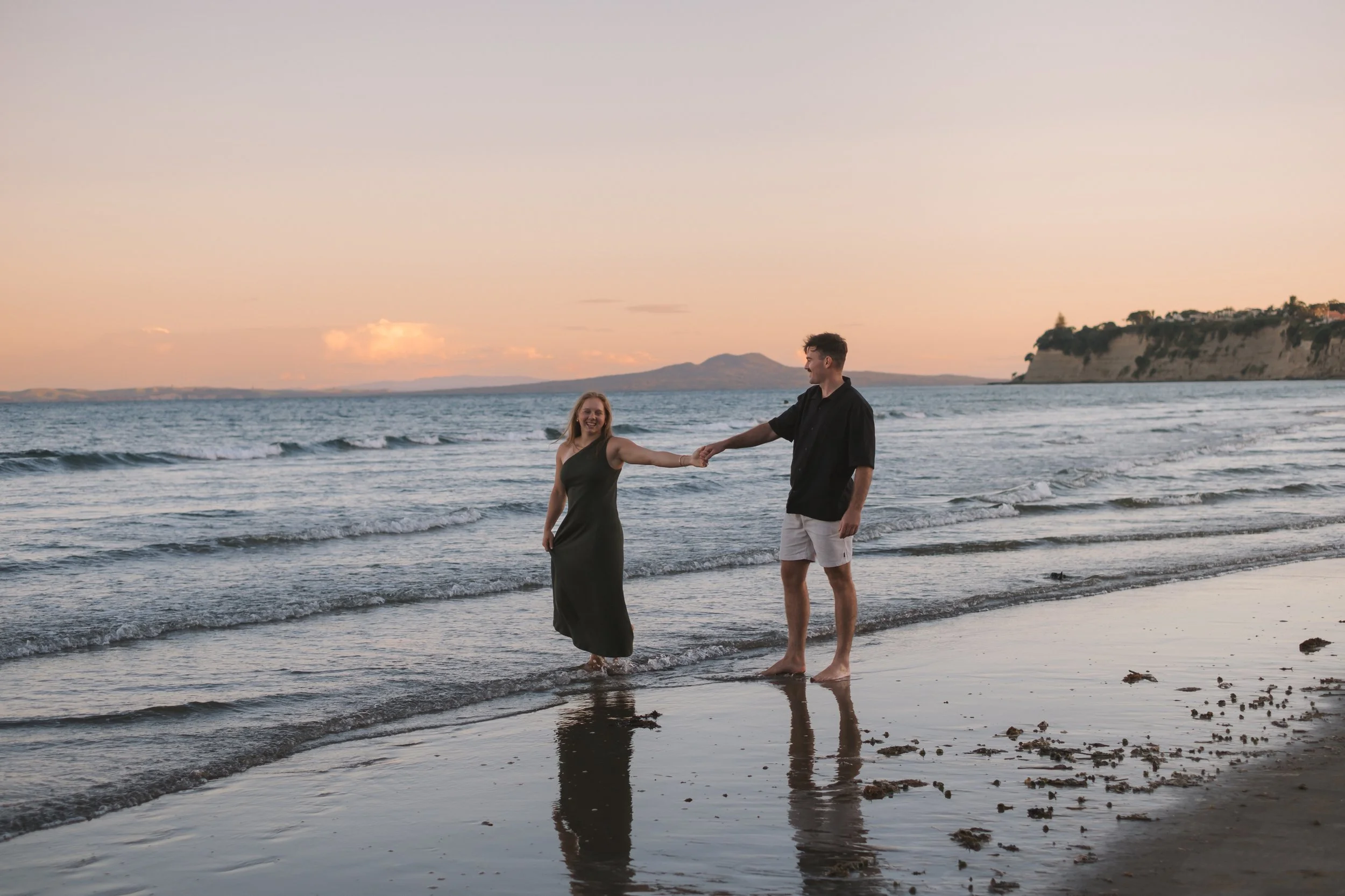 A couple holding hands and smiling at each other while standing in shallow ocean water at sunset, with cliffs and a hill in the background.