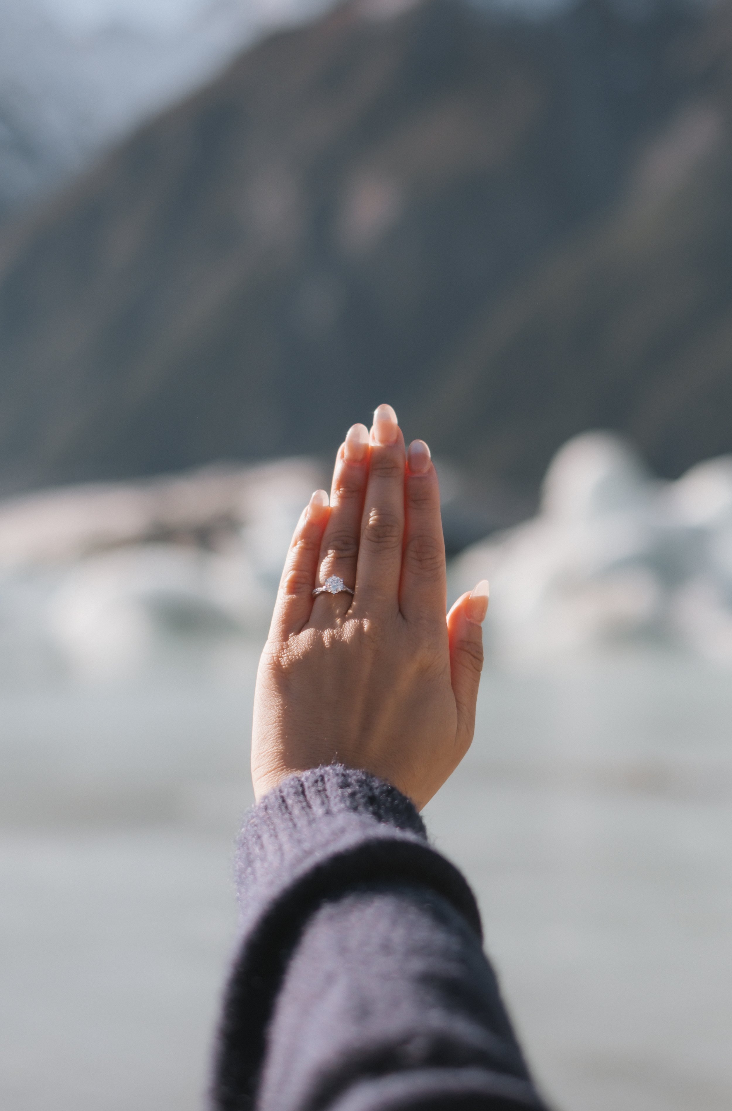 A person's hand is raised in front of a body of water with mountains in the background, wearing a dark sweater with purple accents and a ring with a large gemstone on the ring finger.