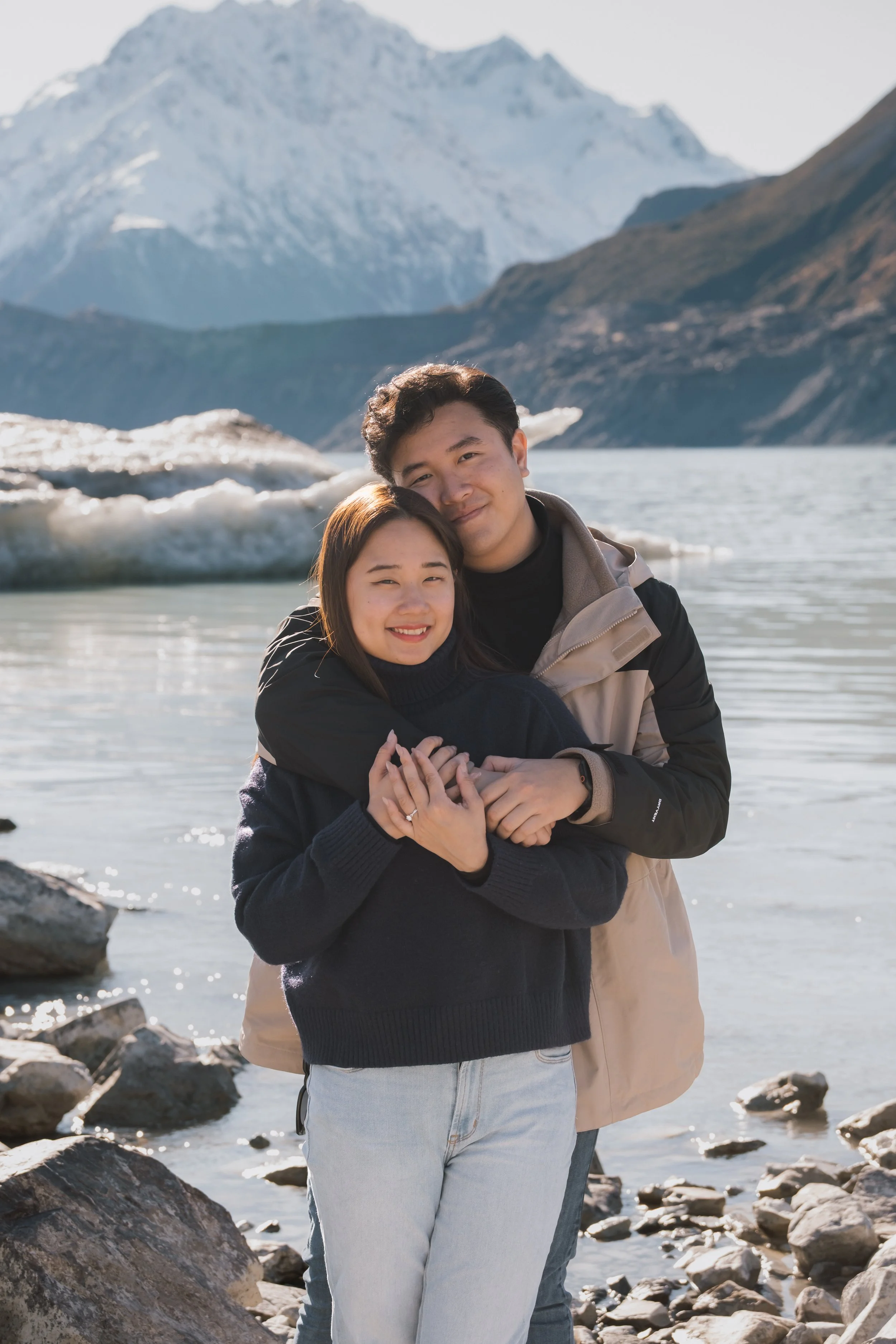 A young couple hugging near a rocky lakeshore with snow-capped mountains in the background.