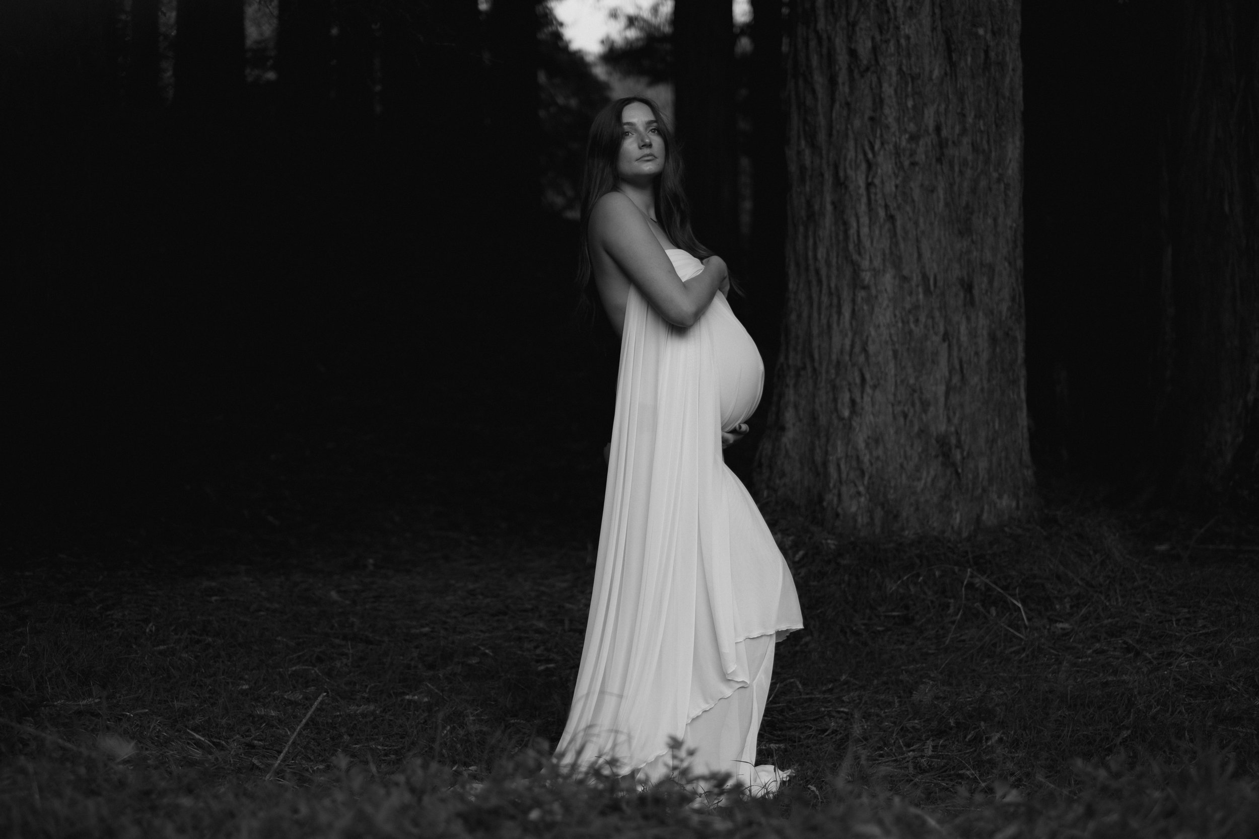 Black and white photo of a pregnant woman in a flowing dress standing in a forest, looking up, with trees in the background.
